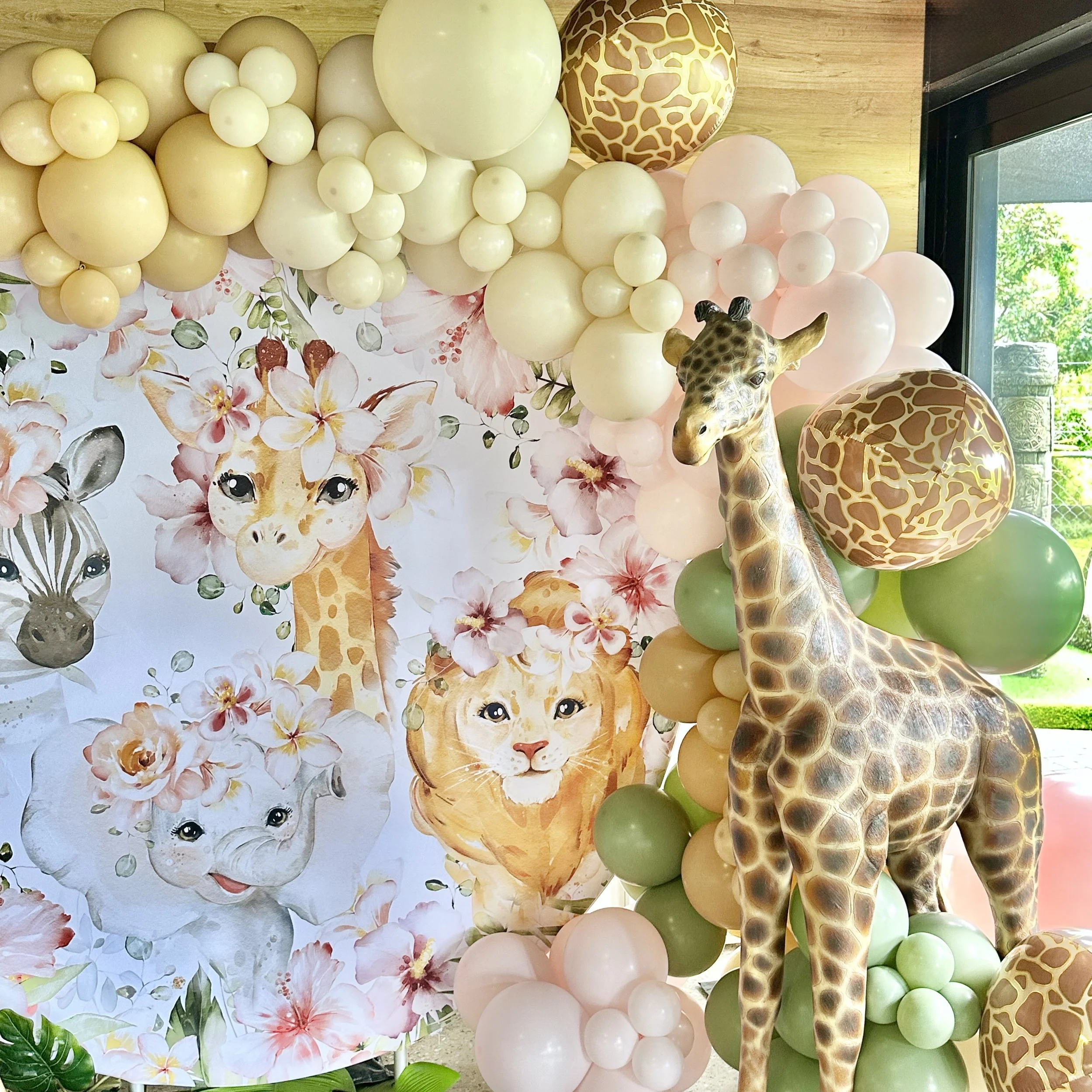Decorative jungle-themed baby shower display with balloons, giraffe sculpture, and illustrated animals including a zebra, lion, elephant, and giraffe surrounded by pink and green floral balloons.