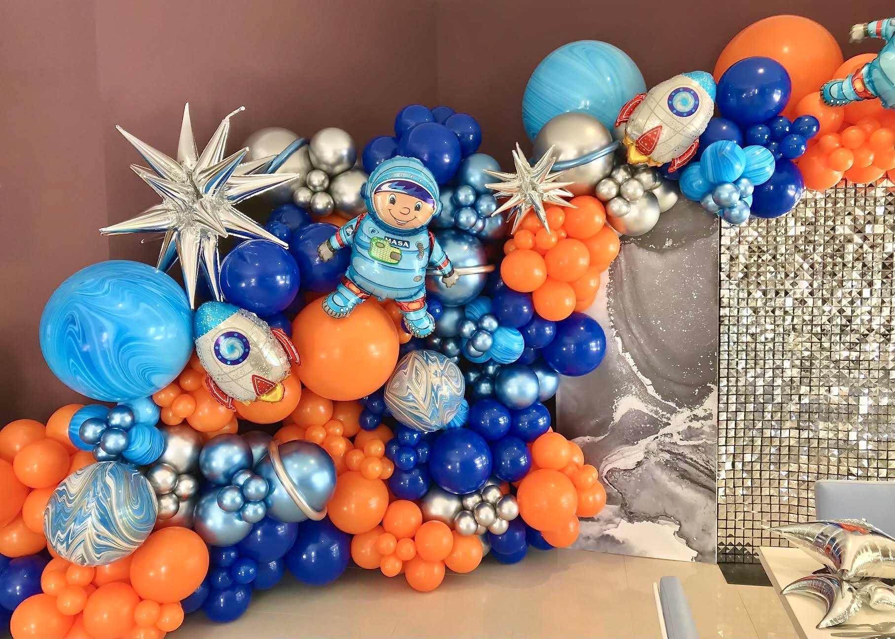 Space-themed balloon decoration with astronaut, rocket, stars, planets, and silver-colored balloons in blue, orange, silver, and metallic tones