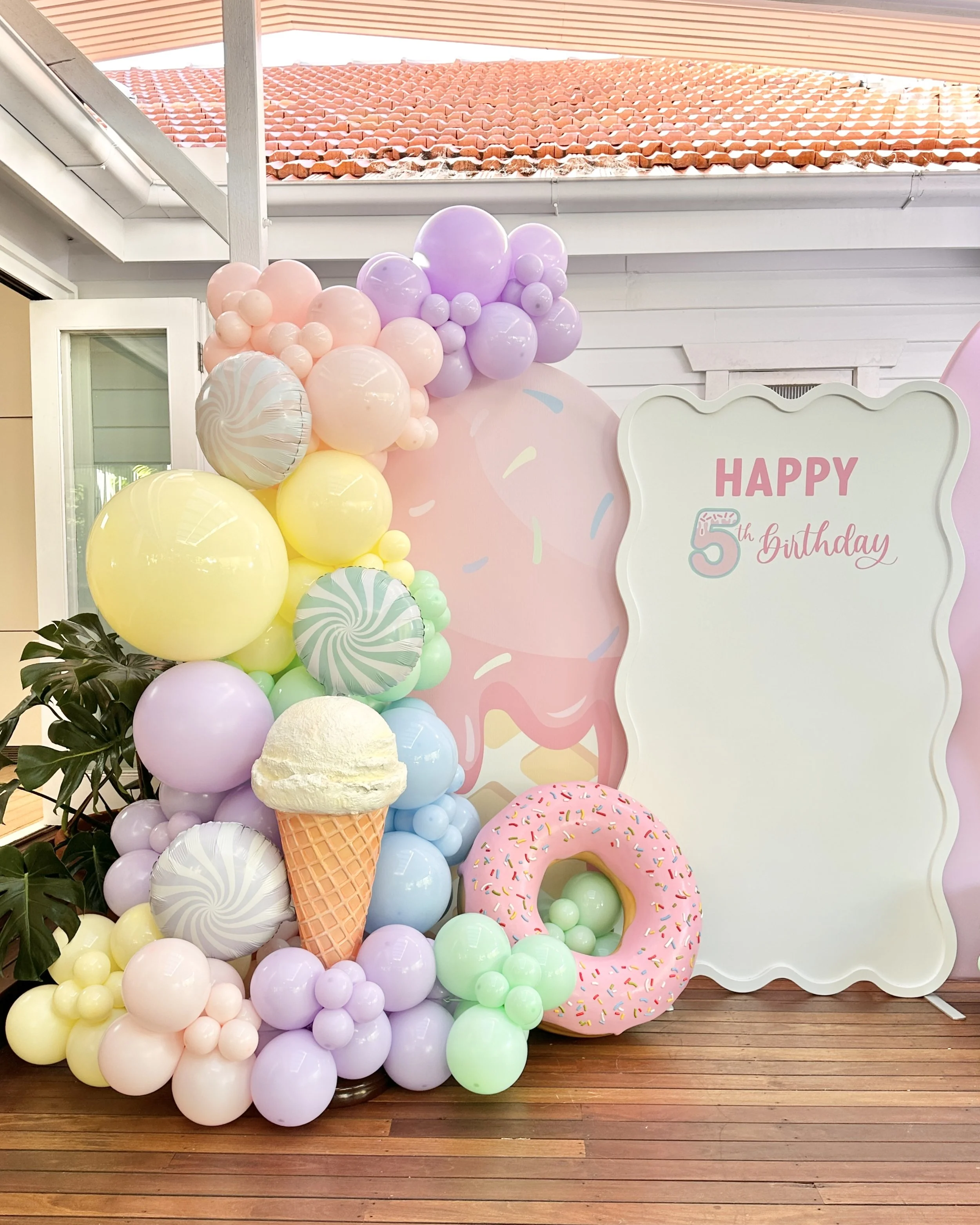 Colorful balloon decorations with ice cream and donut themed props at a birthday party, including a large sign that says "Happy 5th Birthday."
