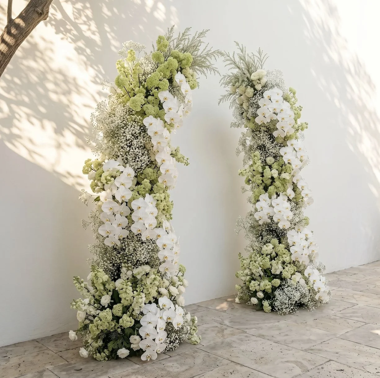 Two large flower arrangements arranged in a semi-circular shape with white and light green flowers, placed outdoors against a white wall with sunlight and shadows.