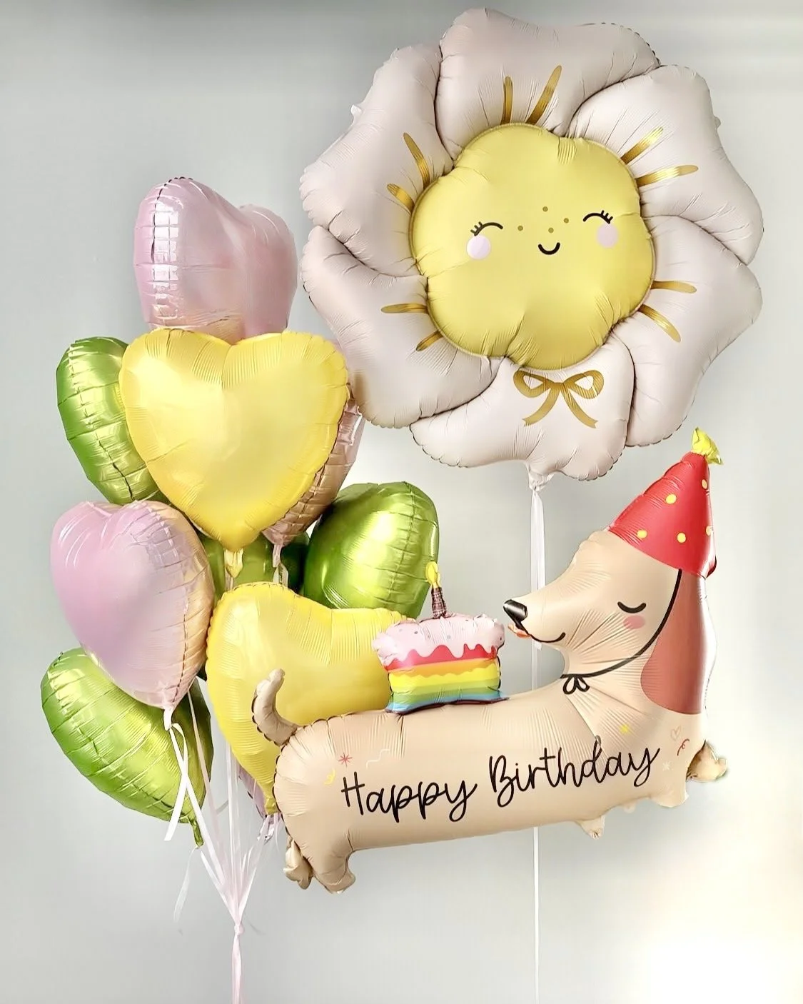 A collection of colorful birthday balloons, including a large smiling sun with a bow, heart-shaped balloons, and a dog with a birthday hat and a birthday cake-shaped balloon with 'Happy Birthday' written on it.