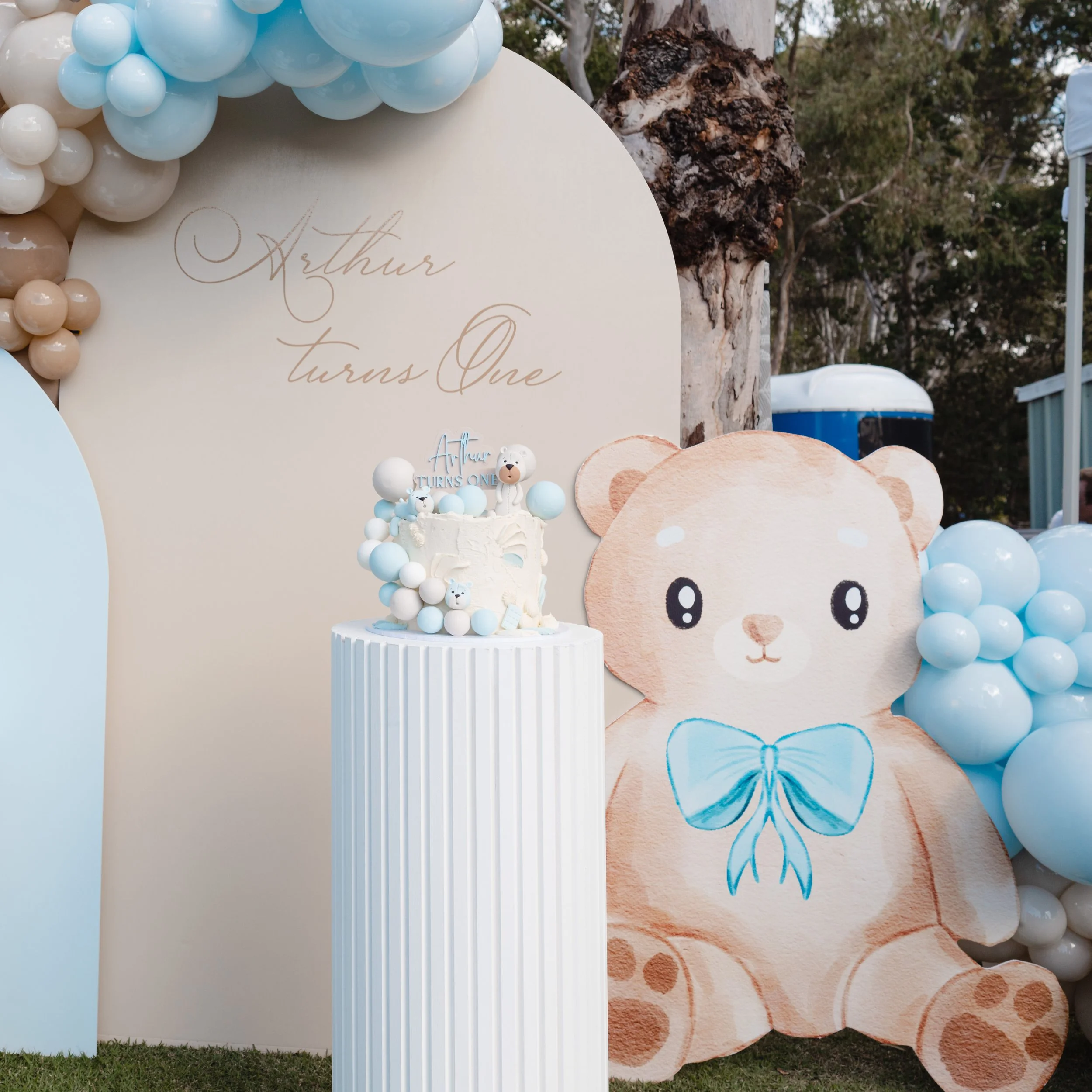 Teddy Bear theme first birthday setup. Includes pastel balloons, a large teddy bear cutout with a blue bow and a balloon backdrop