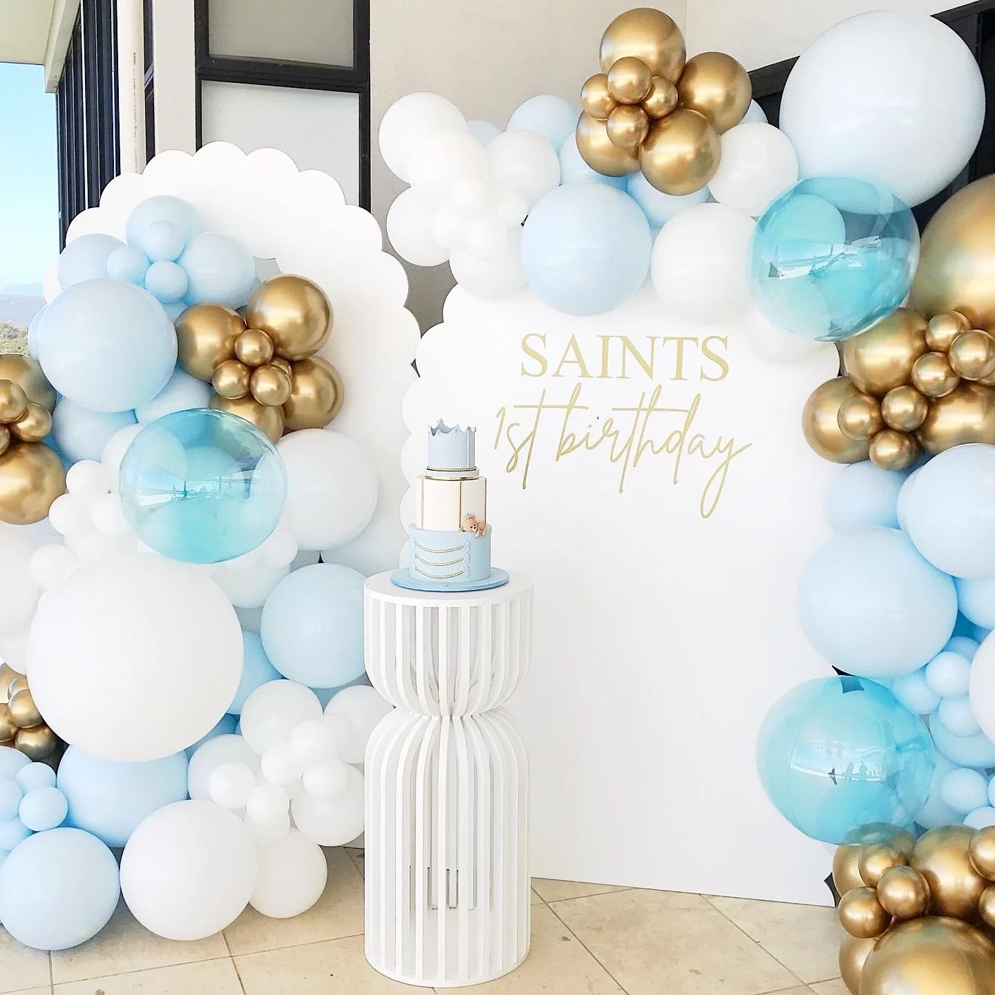 Decorative backdrop with balloons in white, blue, and gold, with a personalised sign for a 1st birthday