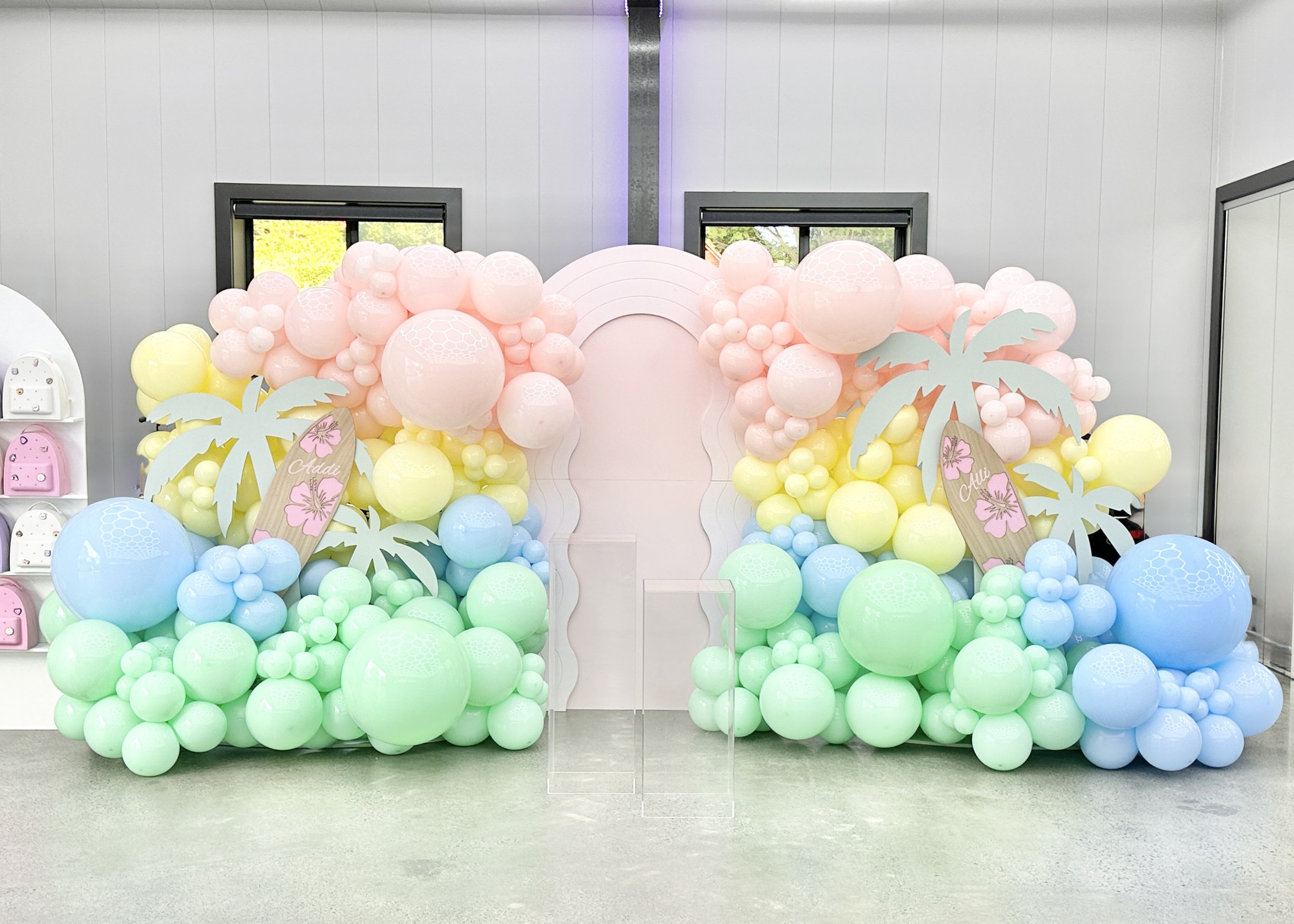 Pastel balloon wall backdrop with palm tree props, custom surfboard signs, for a tropical pool party themed event