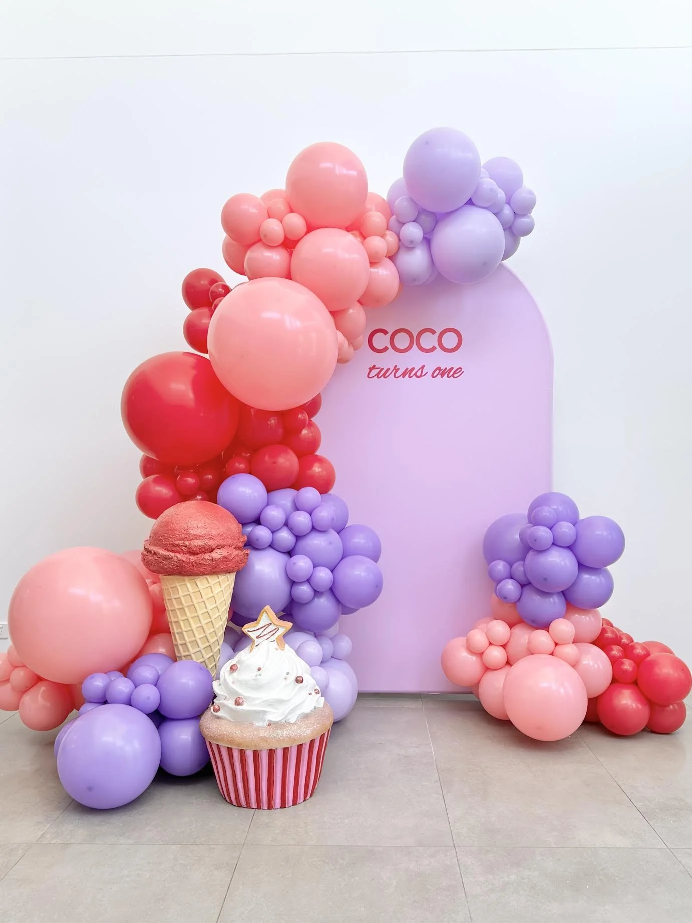 Decorative backdrop with pink and purple balloons, a pink sign that reads "COCO turns one," a large cupcake with pink frosting and white icing with red and pink sprinkles, and a cone-shaped ice cream decoration. The setup suggests a birthday celebrat