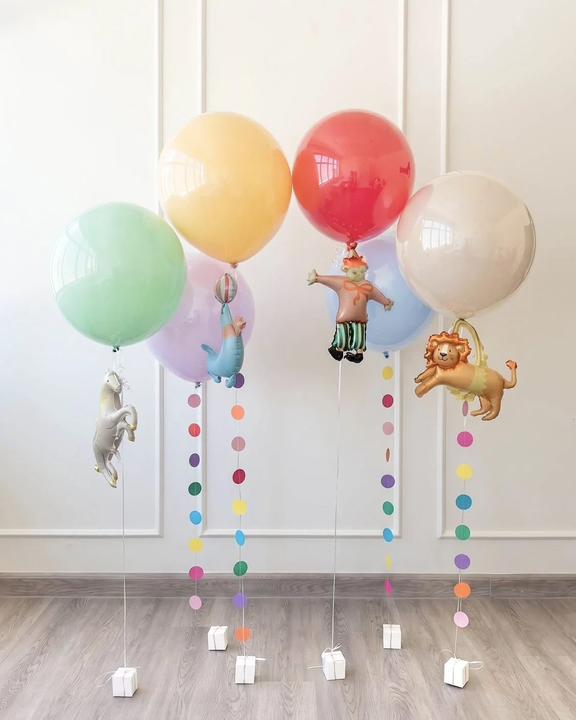 Colorful balloons with animal and clown balloon figures attached, tied to white weight blocks, set against a white paneled wall.