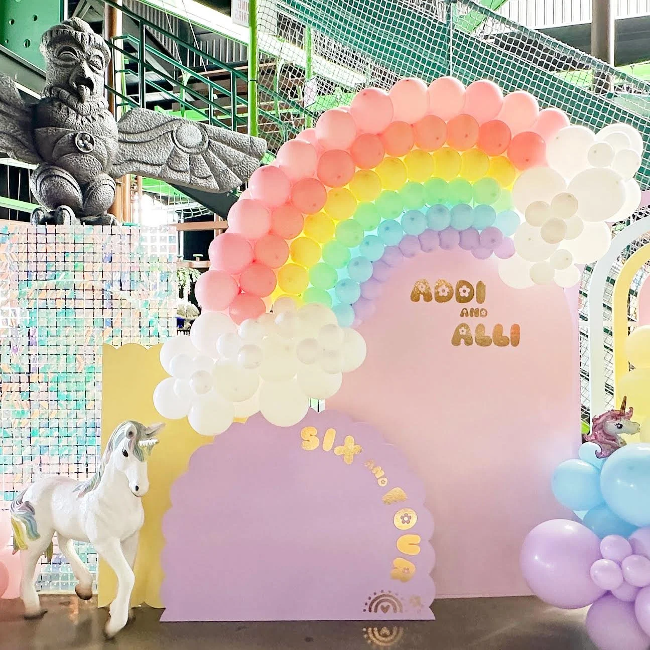 Decorative backdrop with pastel balloons arranged in a rainbow pattern, featuring unicorn sculptures, and a sign that reads "Aodi and Alli".