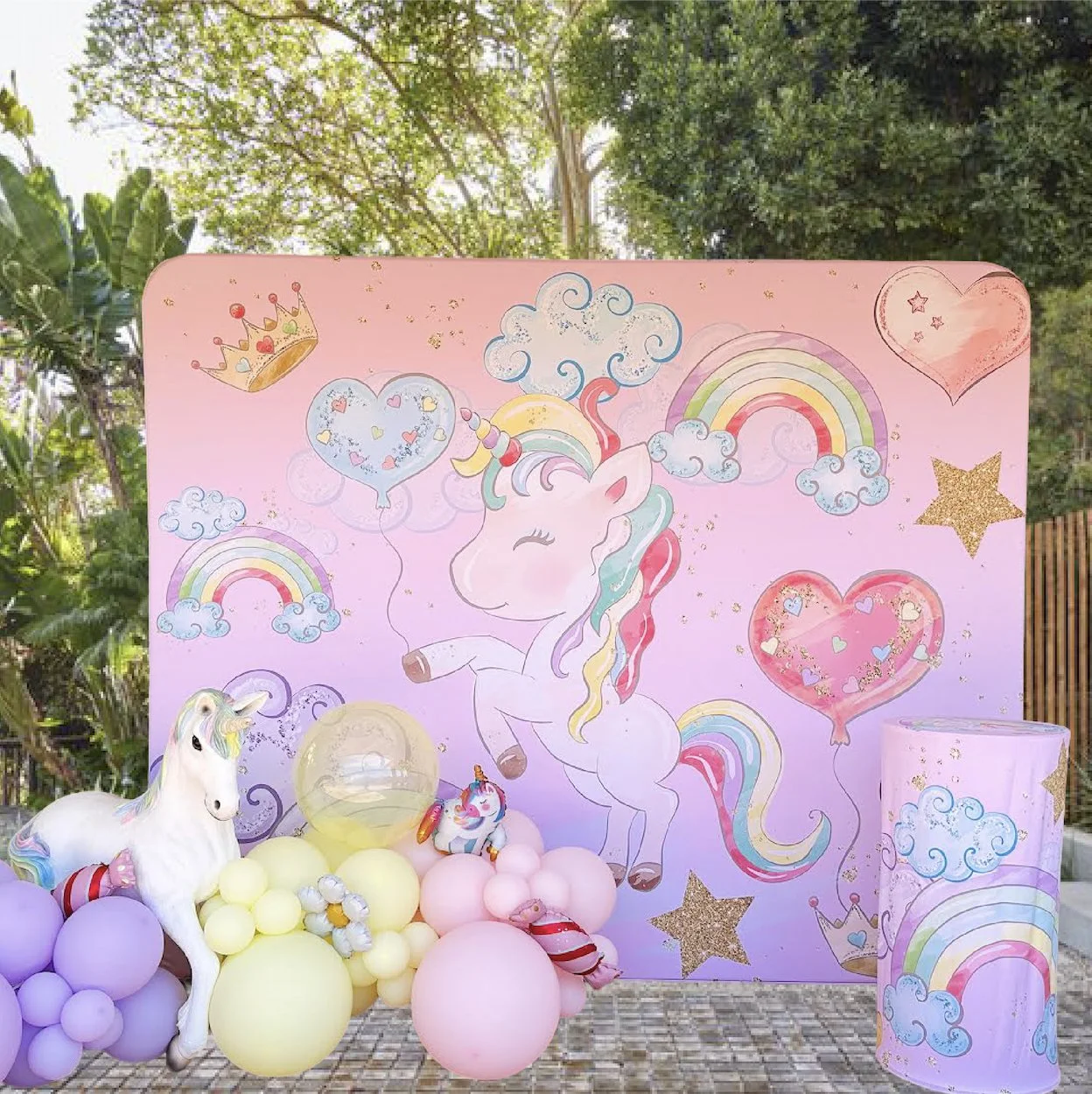 Decorative unicorn-themed setup with pastel-colored balloons, a unicorn prop, and a backdrop featuring a smiling unicorn, rainbows, clouds, hearts, stars, and a crown during daytime outdoors.