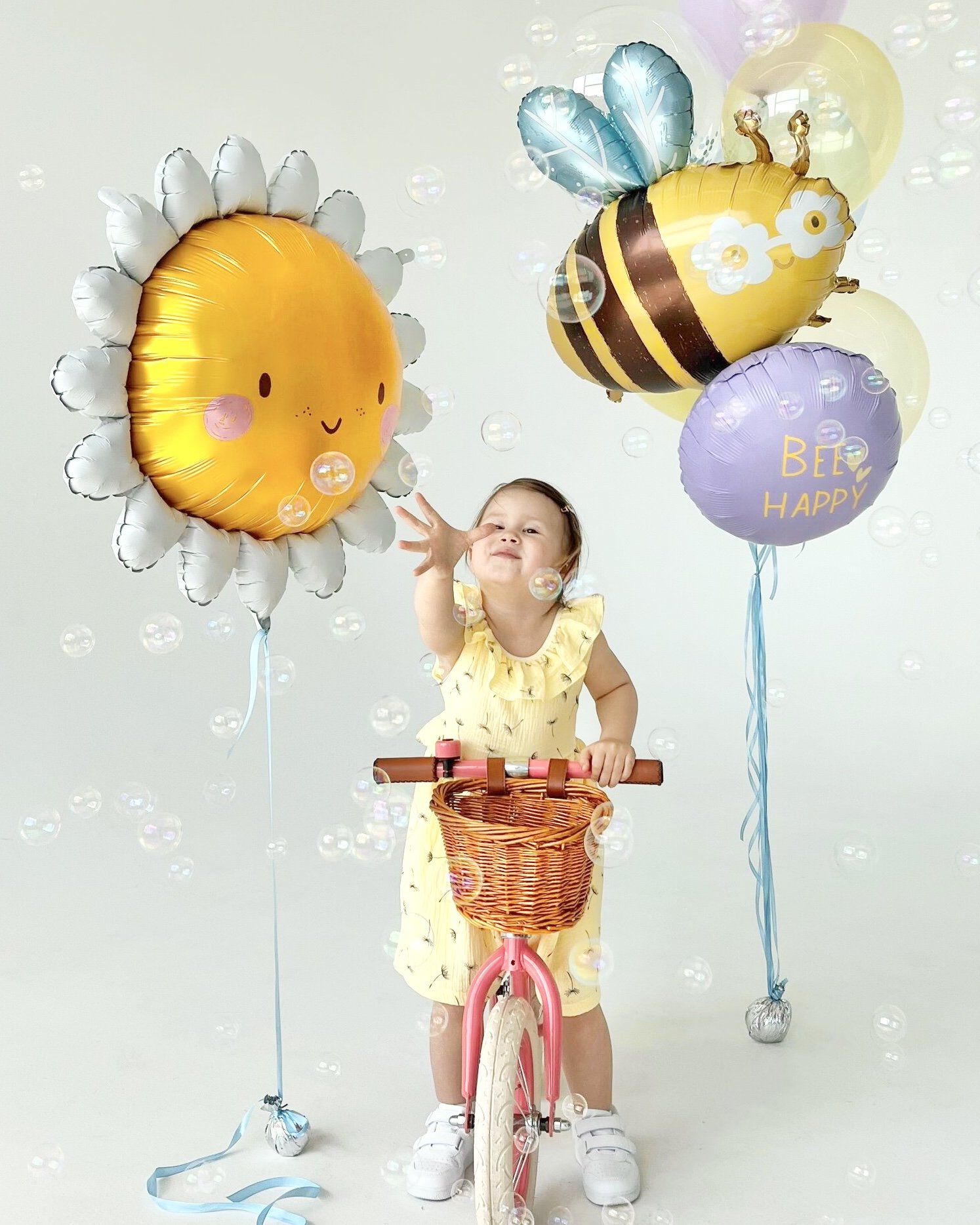 A young girl in a yellow dress and white sneakers is riding a pink and white bicycle. She is surrounded by colorful balloons, including a sunflower-shaped balloon, a bee-shaped balloon with the text "BEE HAPPY," and other balloons. Bubbles are floati