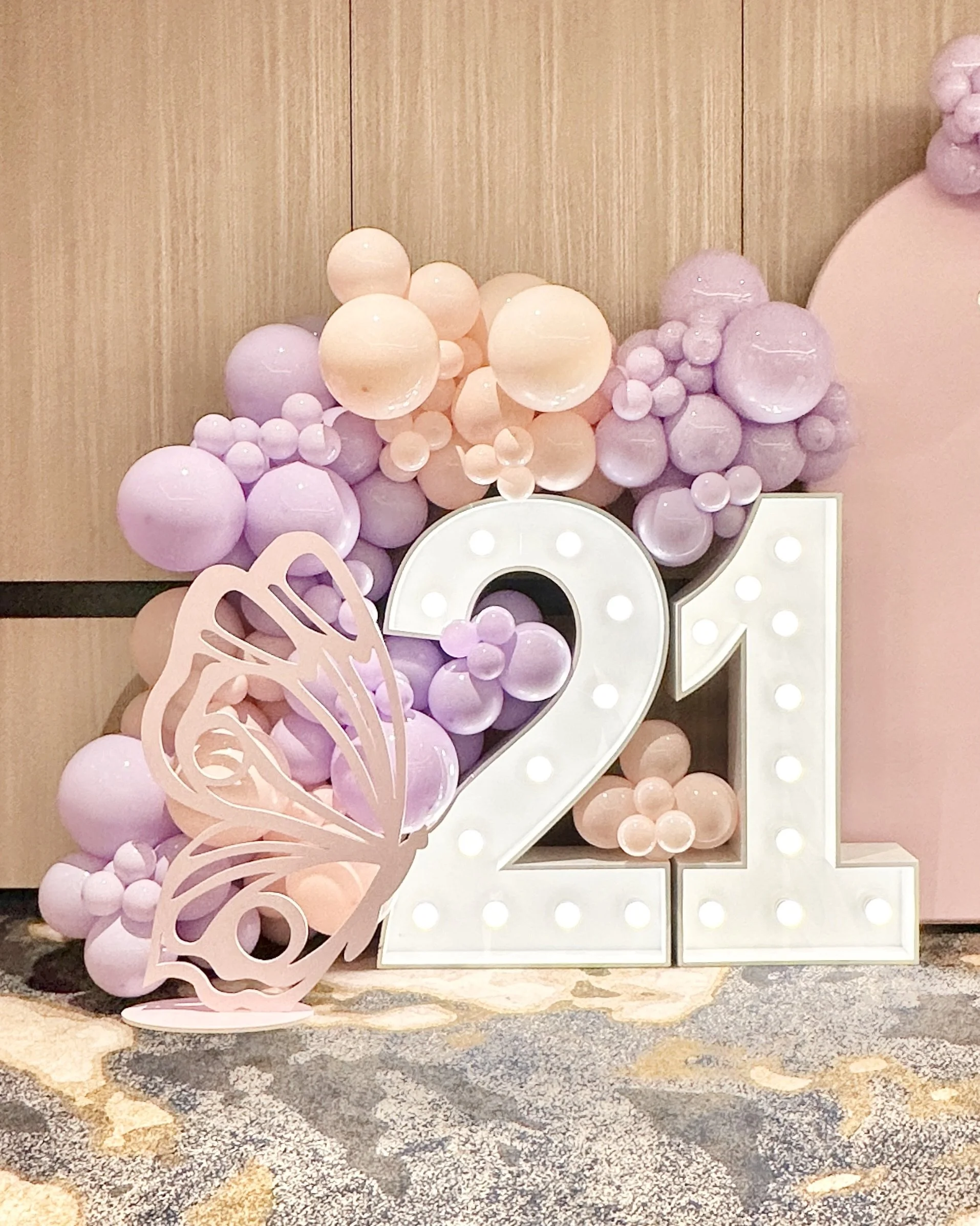 Decorative display with large white illuminated number 21,  pastel pink and lilac balloon wall and a butterfly prop