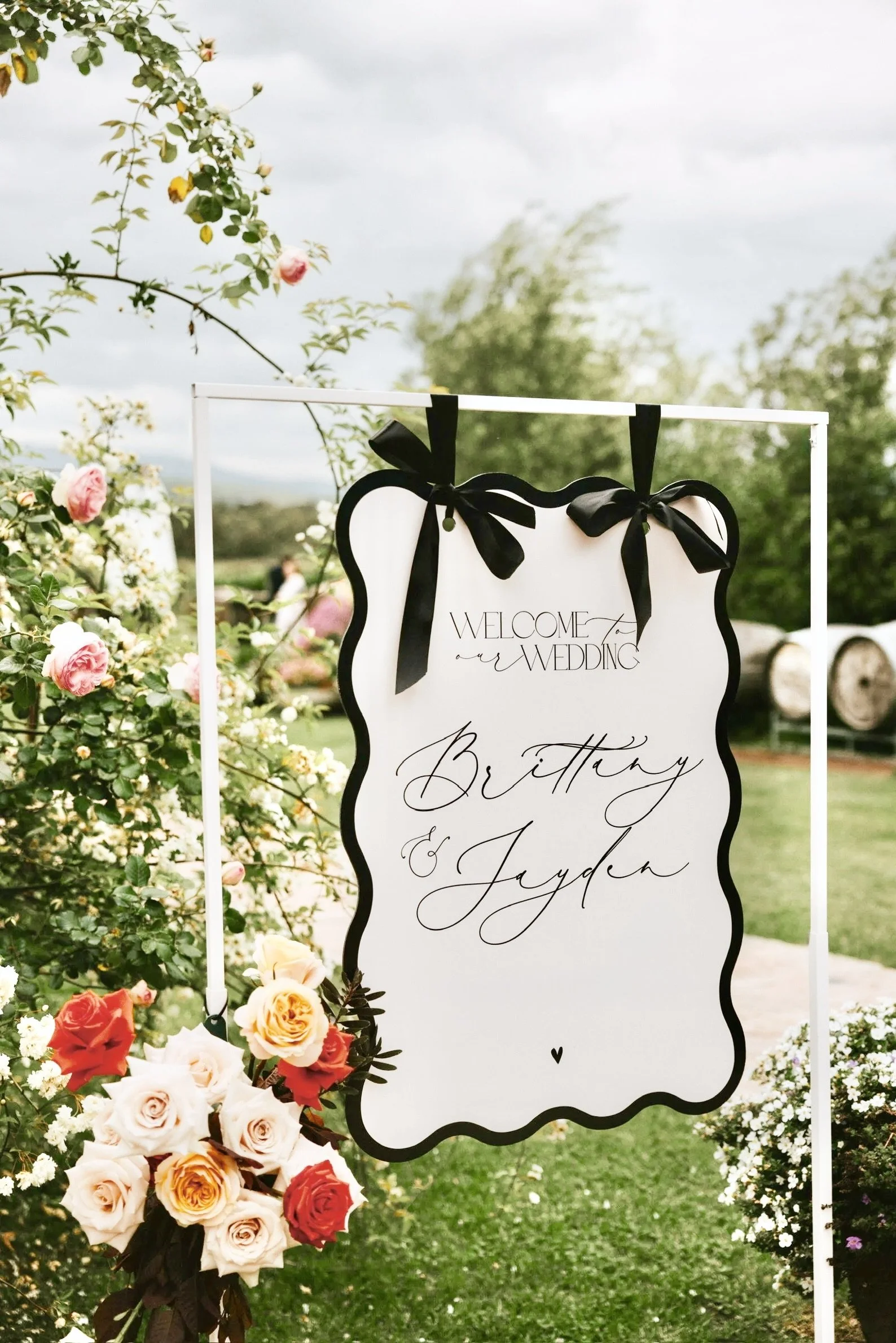 Wedding welcome sign with black ribbon bows, surrounded by colorful roses and flowers in an outdoor garden setting
