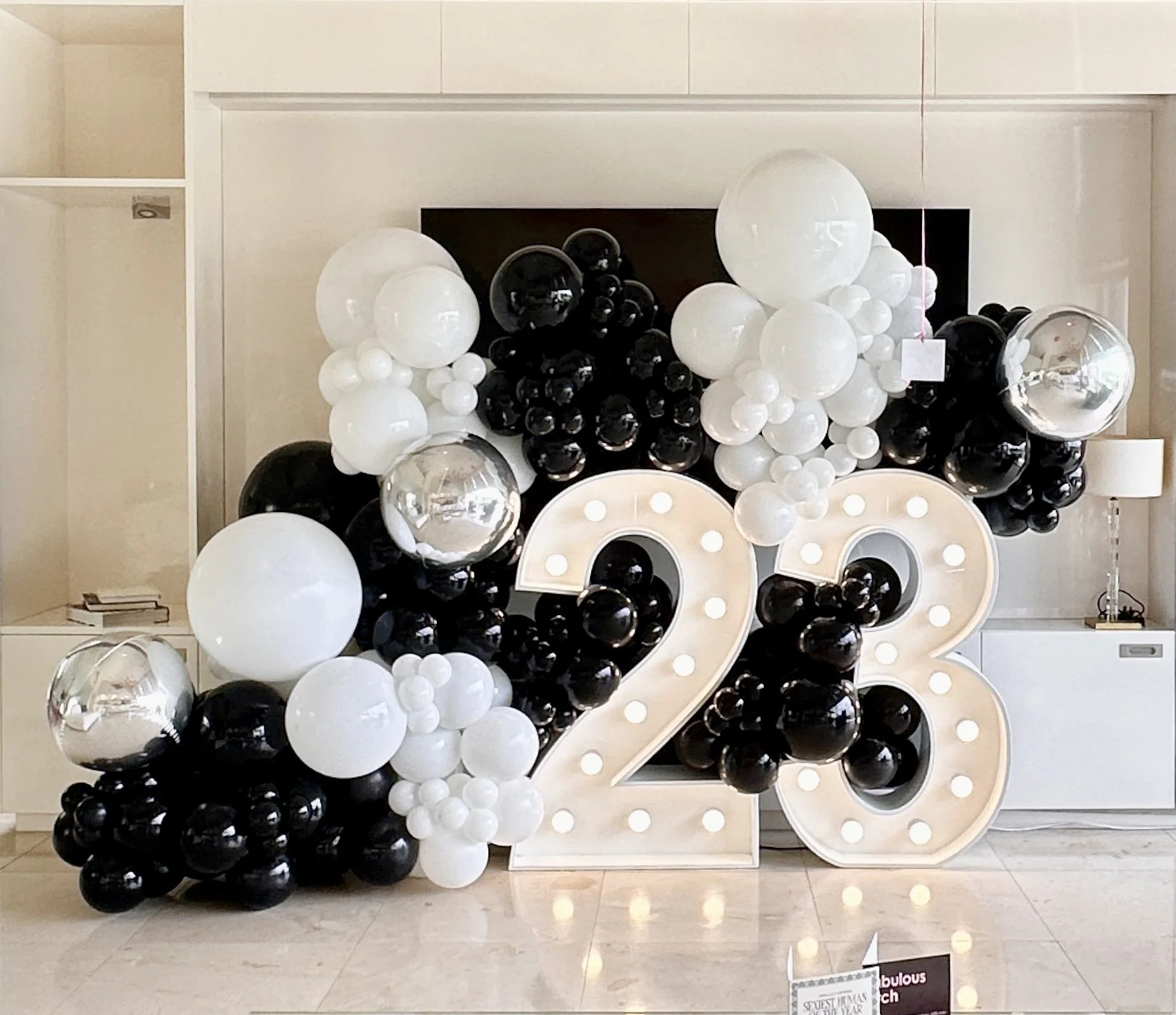 Large decorative balloon display with black, white, and metallic balloons, featuring prominent marquee lights forming the numbers 23