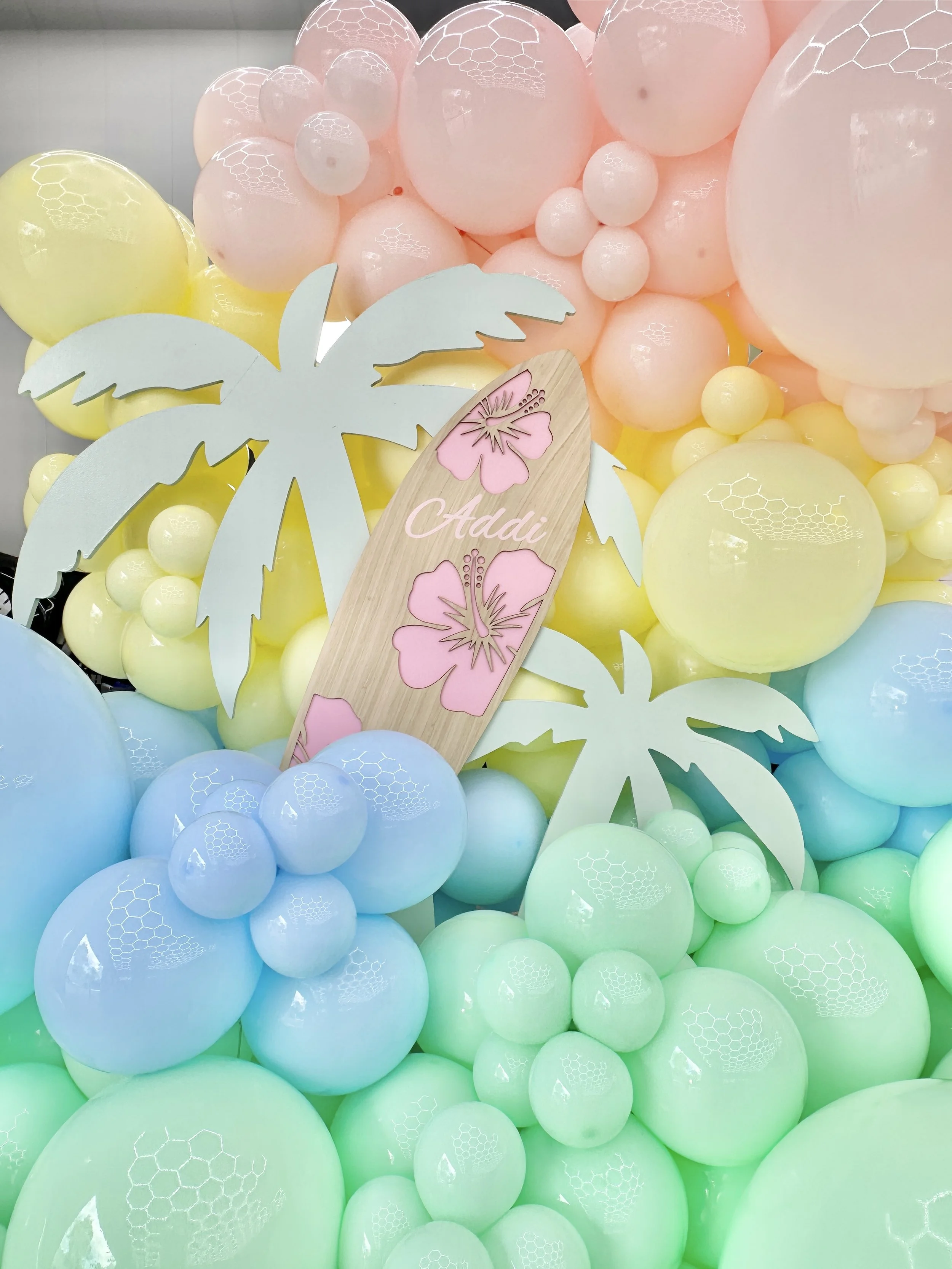 Pastel balloon wall around palm tree props and custom surfboard signs, for a tropical pool party themed event