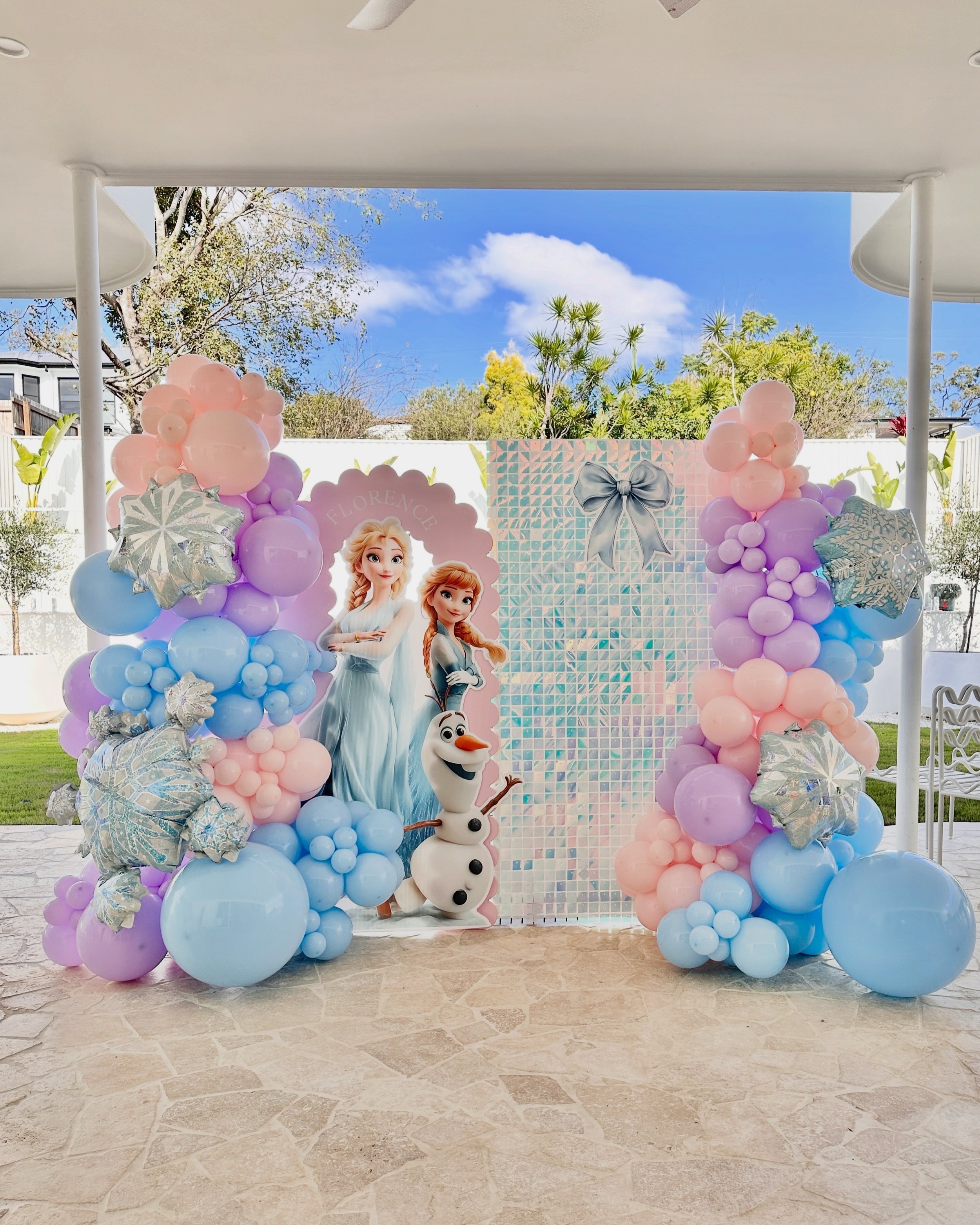 Decorated backdrop with balloons in pastel pink, blue, and purple, featuring characters from 'Frozen' including Elsa, Anna, and Olaf, for a children's birthday party.