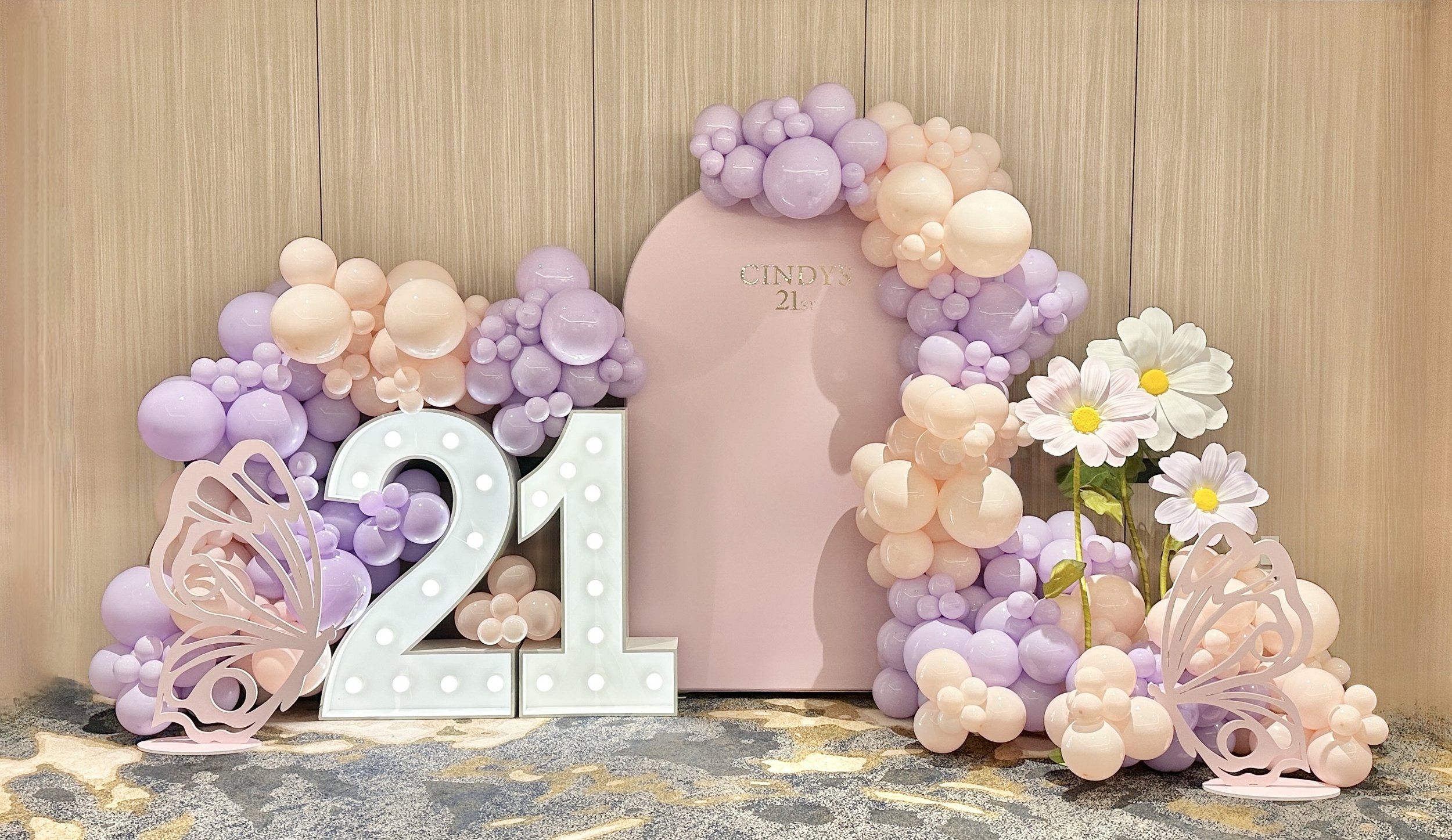 Decorative birthday celebration setup featuring a large pink number '21' with white lights, surrounded by pastel purple and peach balloons, large pink and white paper butterflies, and white daisies with yellow centers against a wooden backdrop.