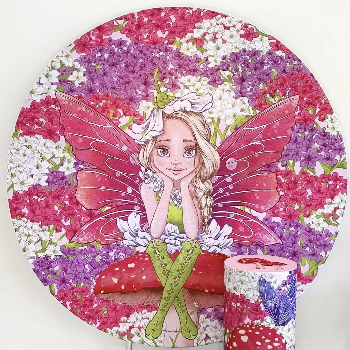 Round fairy backdrop. Fairy with pink wings and blonde hair, sitting with hands on her cheeks, surrounded by pink, purple, and white flowers, with a matching cupcake featuring a blue butterfly and red mushroom in front