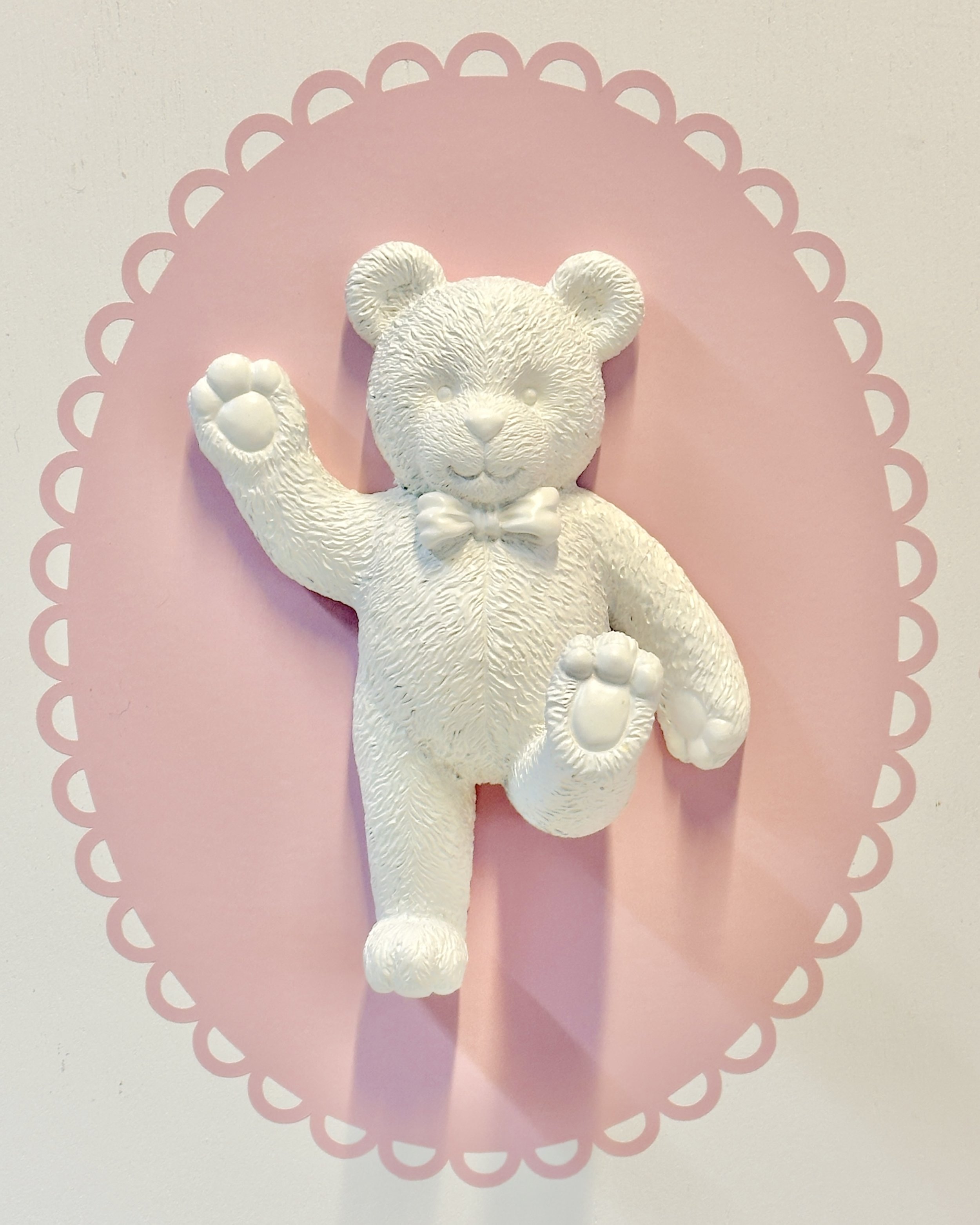White teddy bear figurine with a bow tie, lying on its back on pink decorative paper with a scalloped edge.