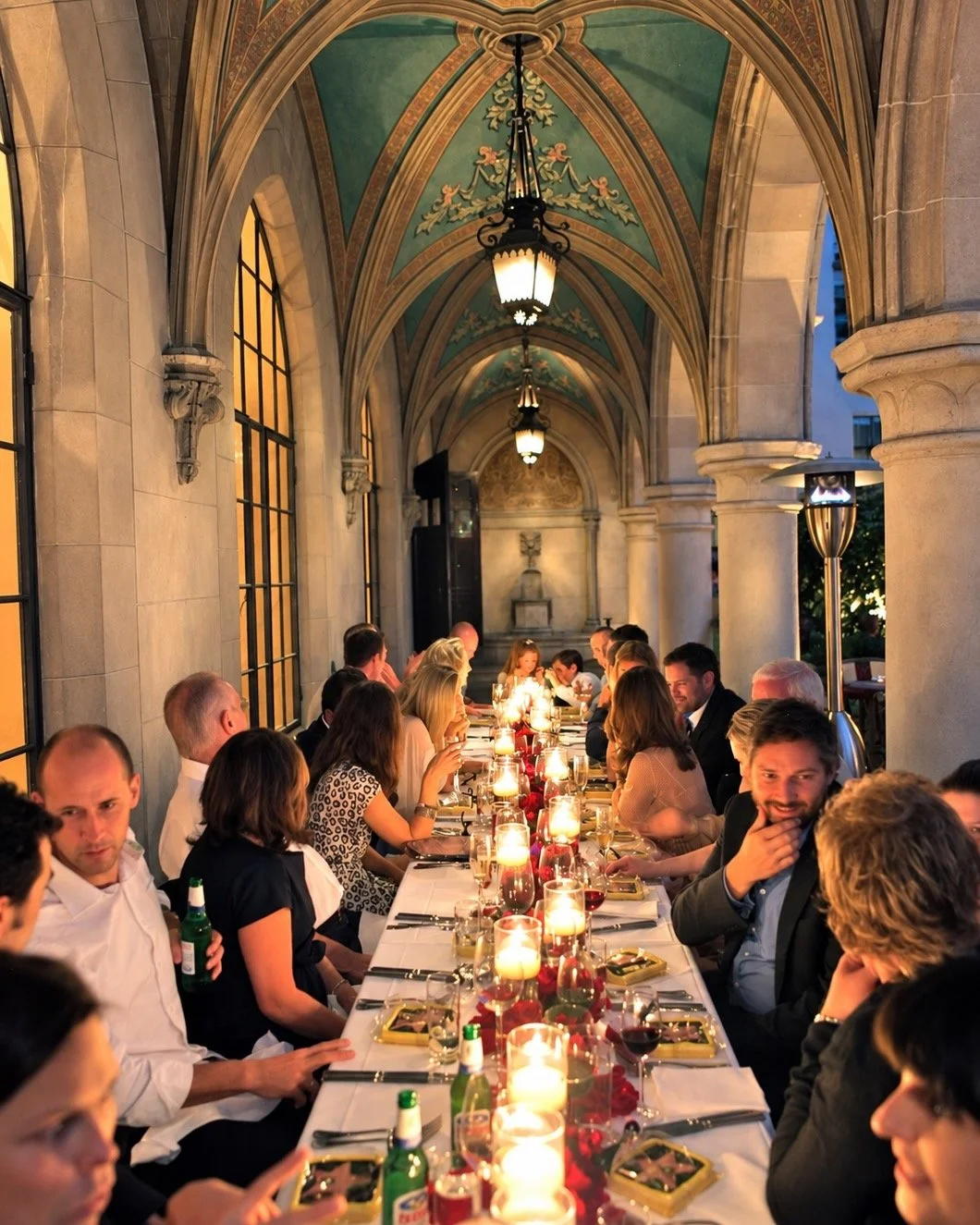 A long, elegantly set dining table with candles, surrounded by well-dressed people in an arched, ornate hall or terrace during evening, with warm lighting and a classical decor backdrop.