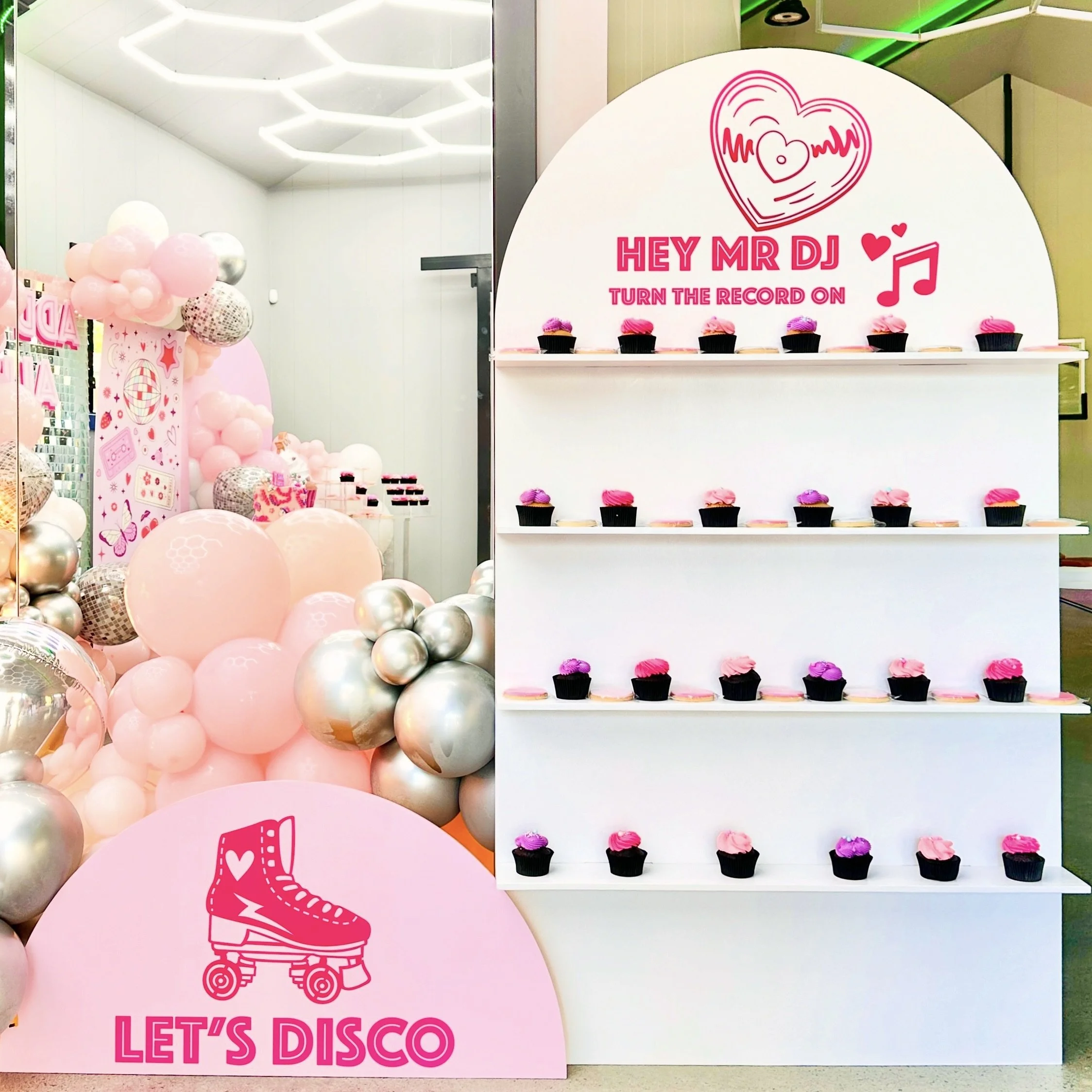 Disco party theme using our Party Favour treat wall with a pink and white roller skate sign reading "Let's Disco" holding themed cupcakes and cookies