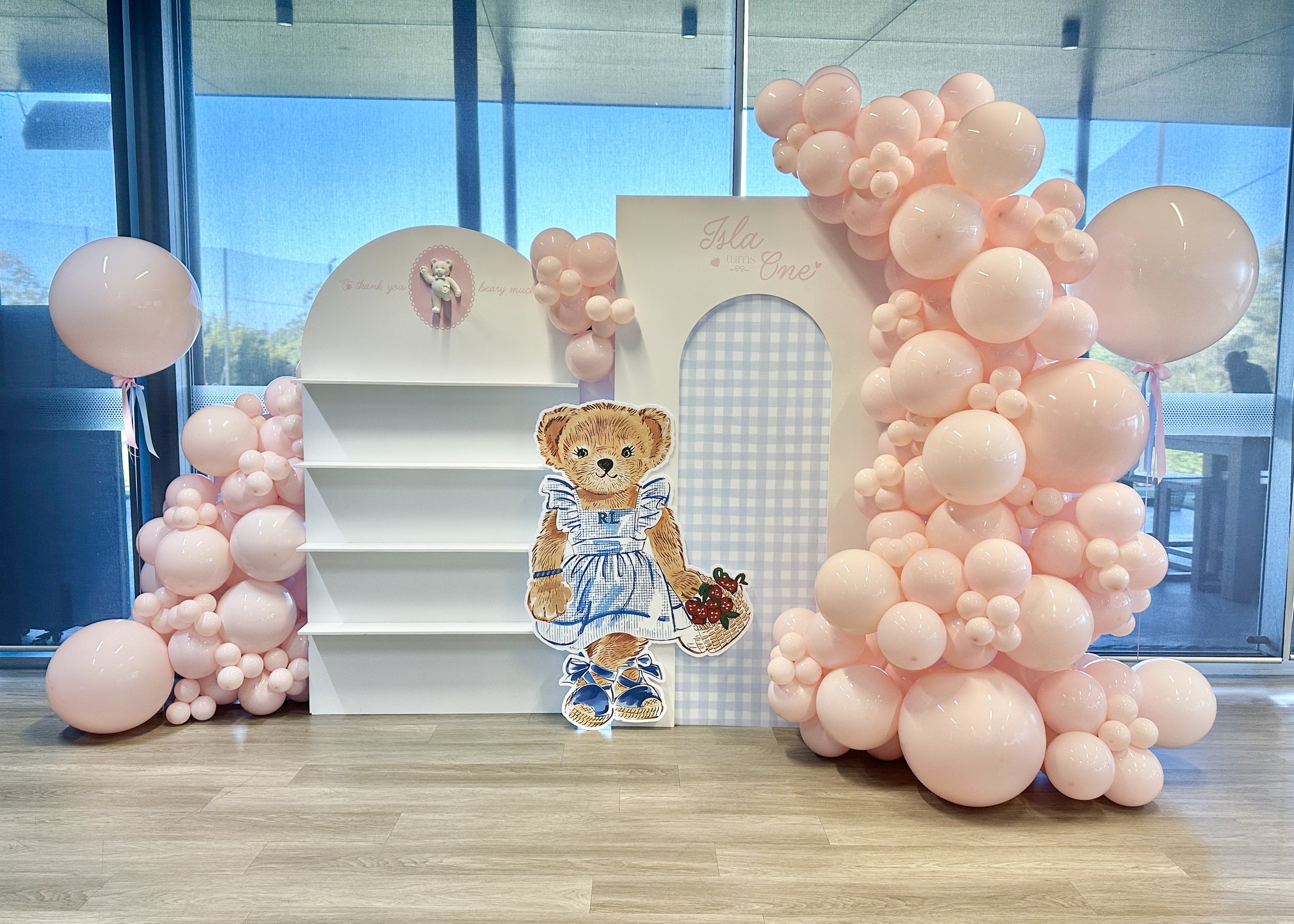 Decorative photo backdrop for a child's first birthday with pink balloons and a white bookshelf, featuring a teddy bear, a birthday card that reads 'Ila turns One', and a cutout of a teddy bear wearing a blue and white striped shirt, set indoors near