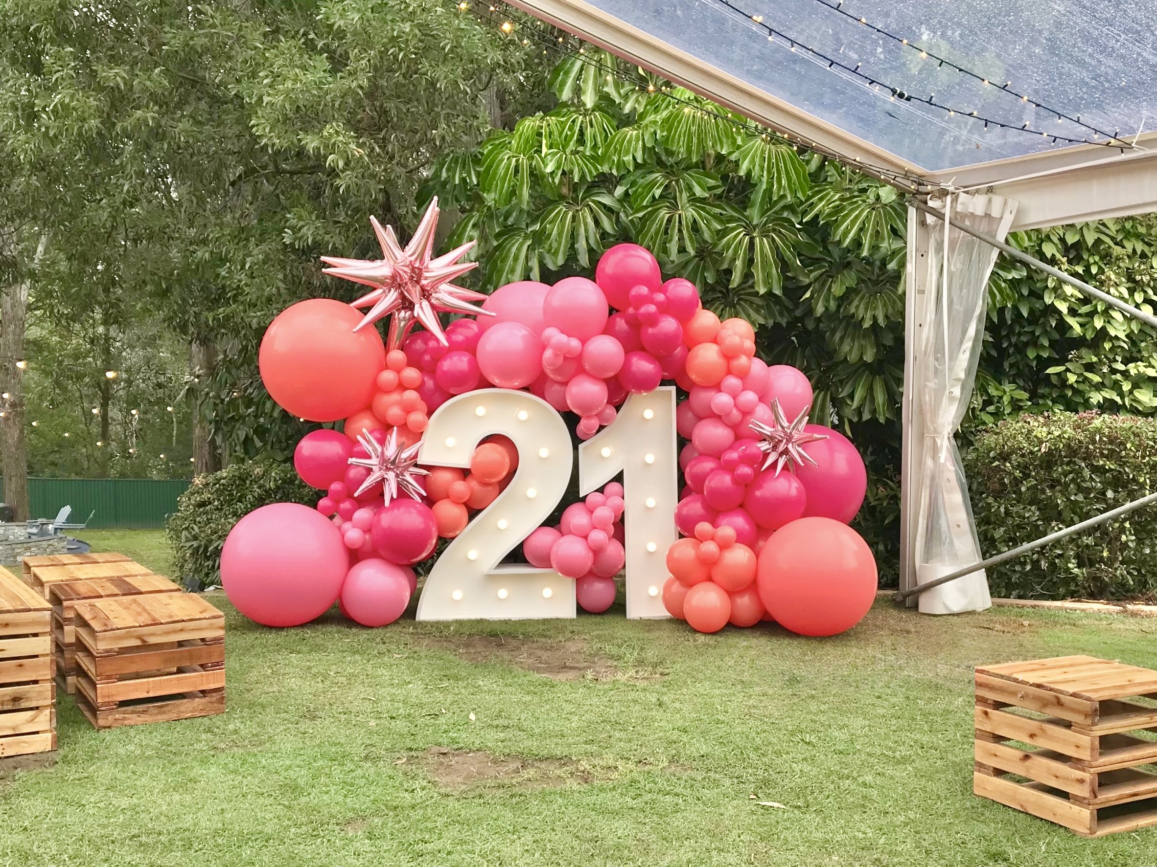 Decorative 2023 birthday celebration setup with pink and coral balloons, white illuminated number 2, greenery background, and string lights under a canopy.