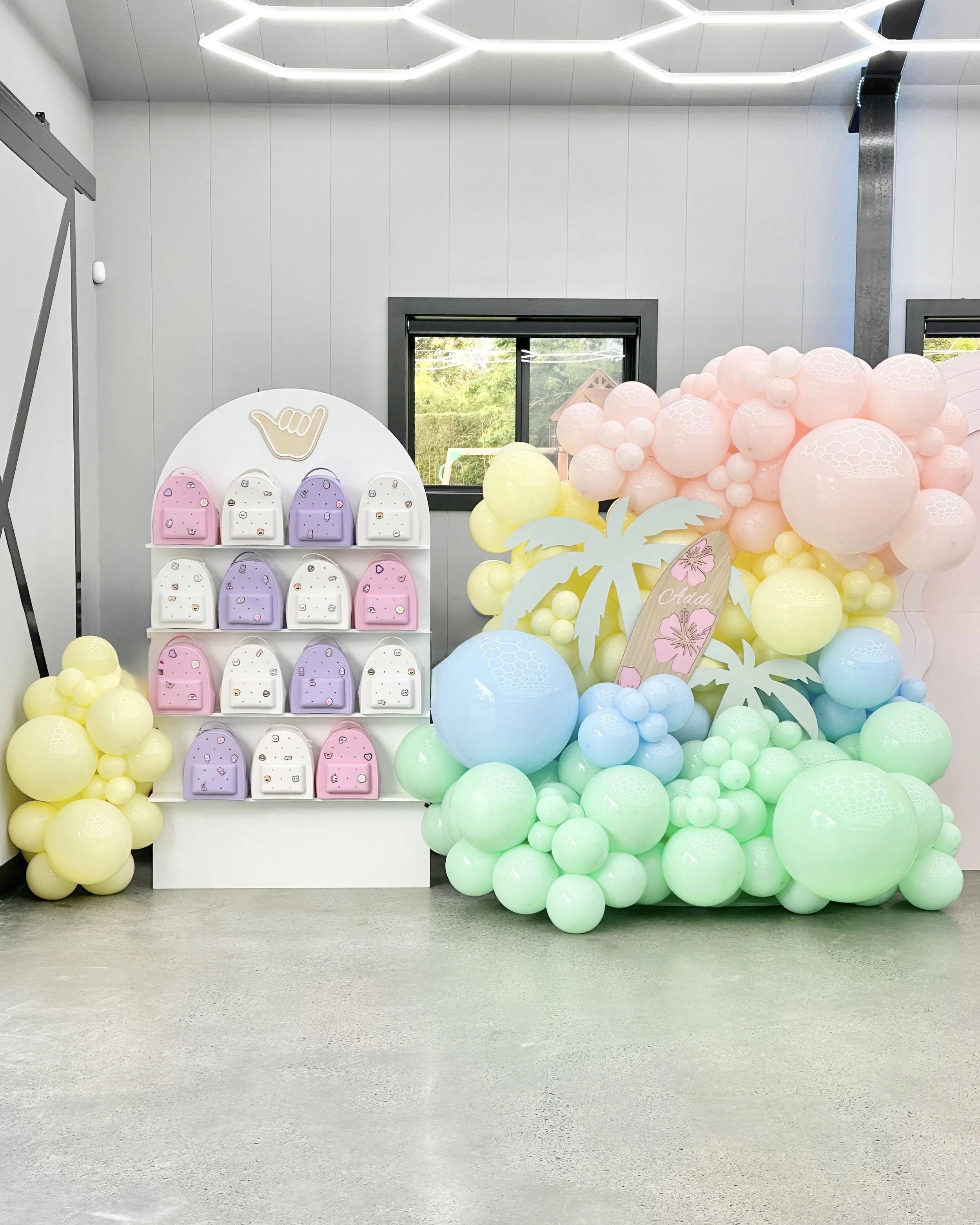 Indoor scene with a display of pastel-colored balloons in shades of yellow, pink, green, and blue, arranged in a cluster. There is also a white, round display stand with small backpacks in pink, white, and purple colors, decorated with small patches.