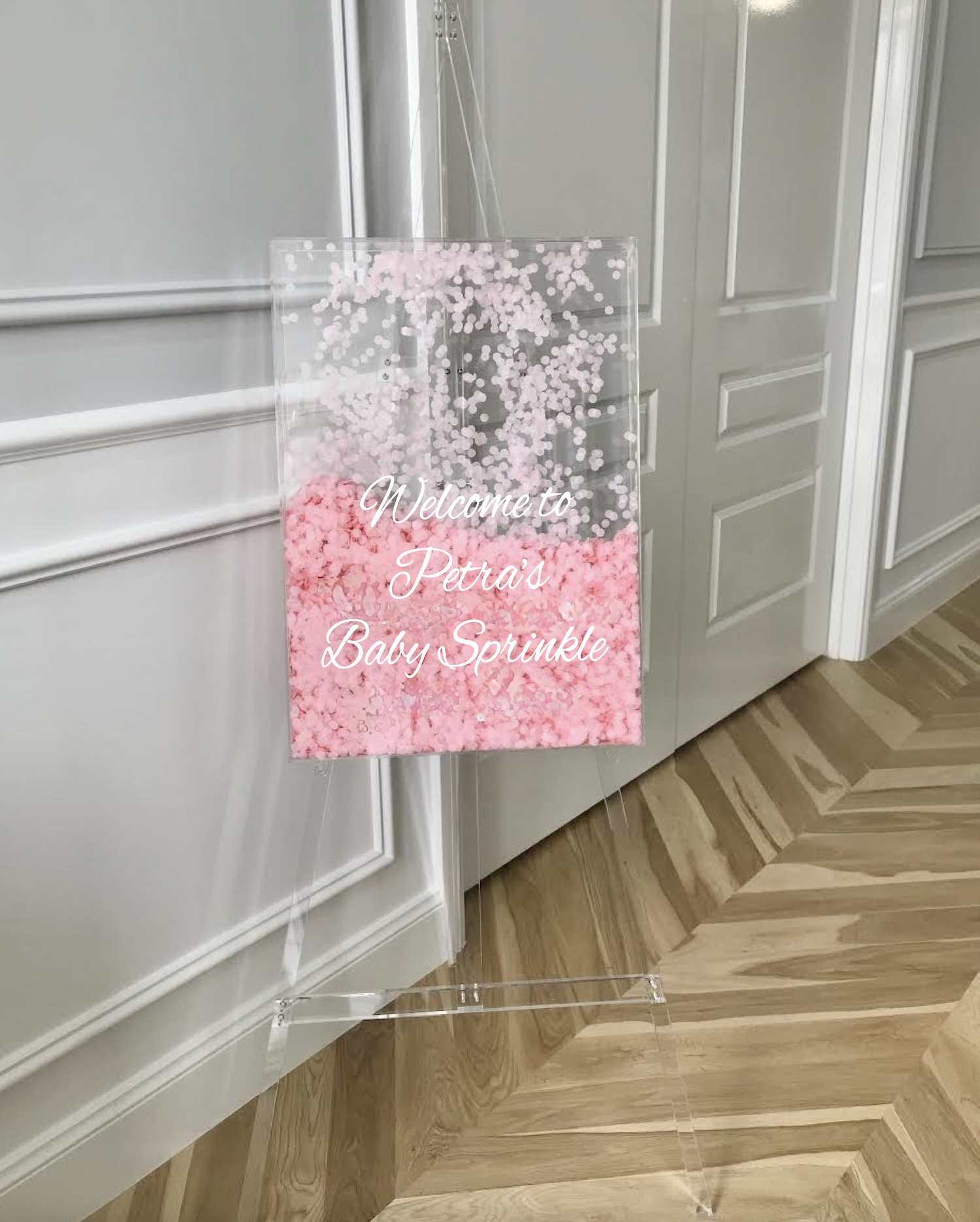 Clear acrylic welcome sign with pink and white confetti inside, featuring the text 'Welcome to Petra's Baby Sprinkle' in white cursive font, leaning against a white wall with paneled doors.