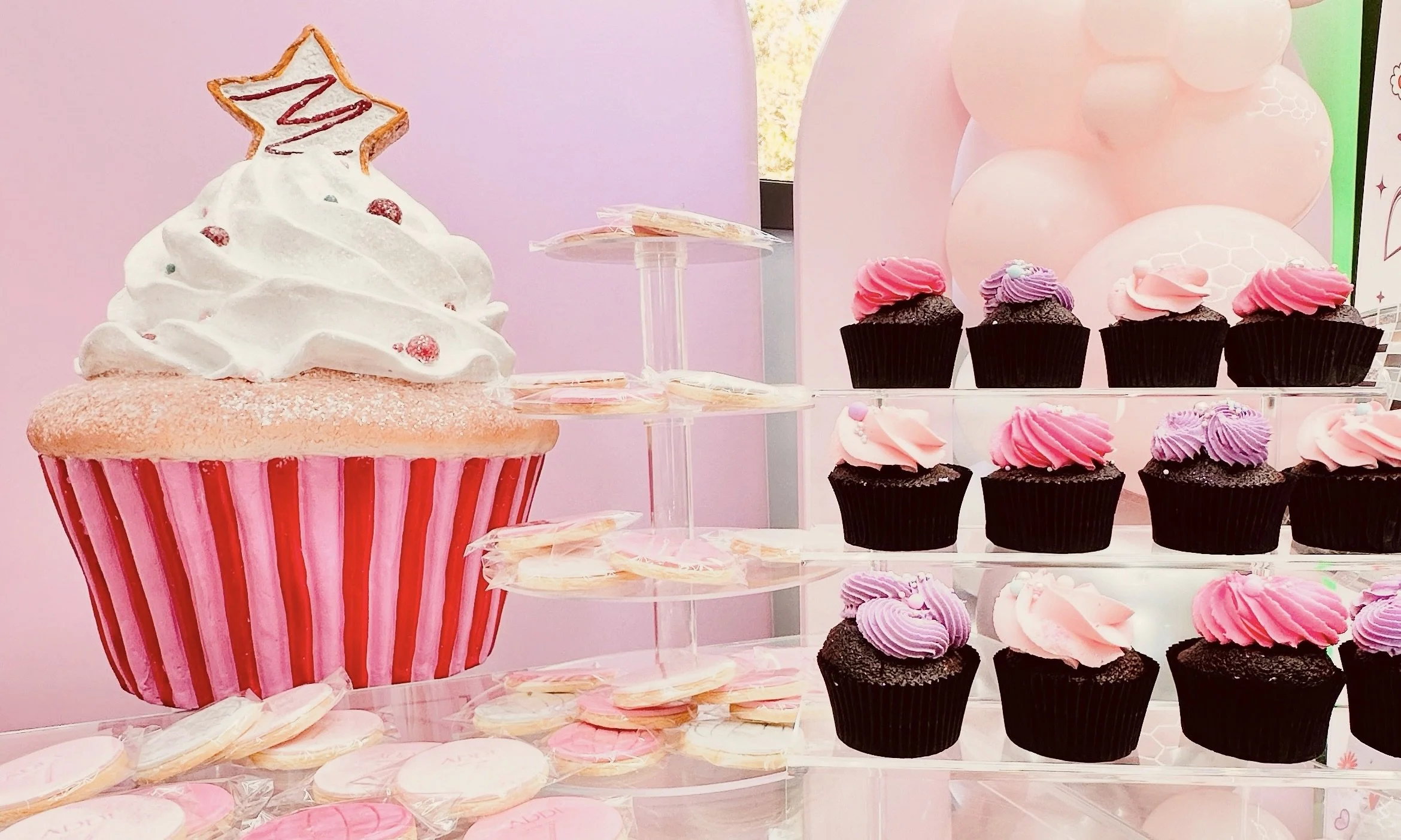 Display of cupcakes and cookies on a clear acrylic dessert table against a pink arch backdrop and a life size cupcake prop