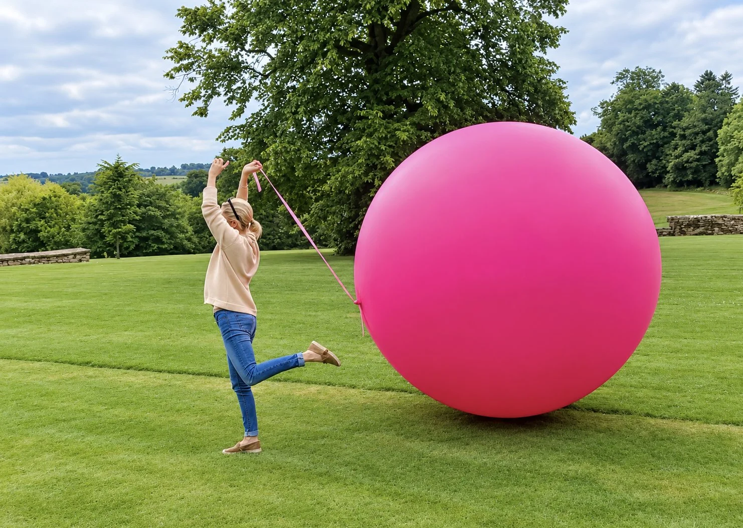A girl with blonde hair tied in a ponytail wearing a beige sweater, blue jeans, and beige shoes holding a pink string attached to a large pink inflatable ball on a grassy field with trees and a cloudy sky in the background.