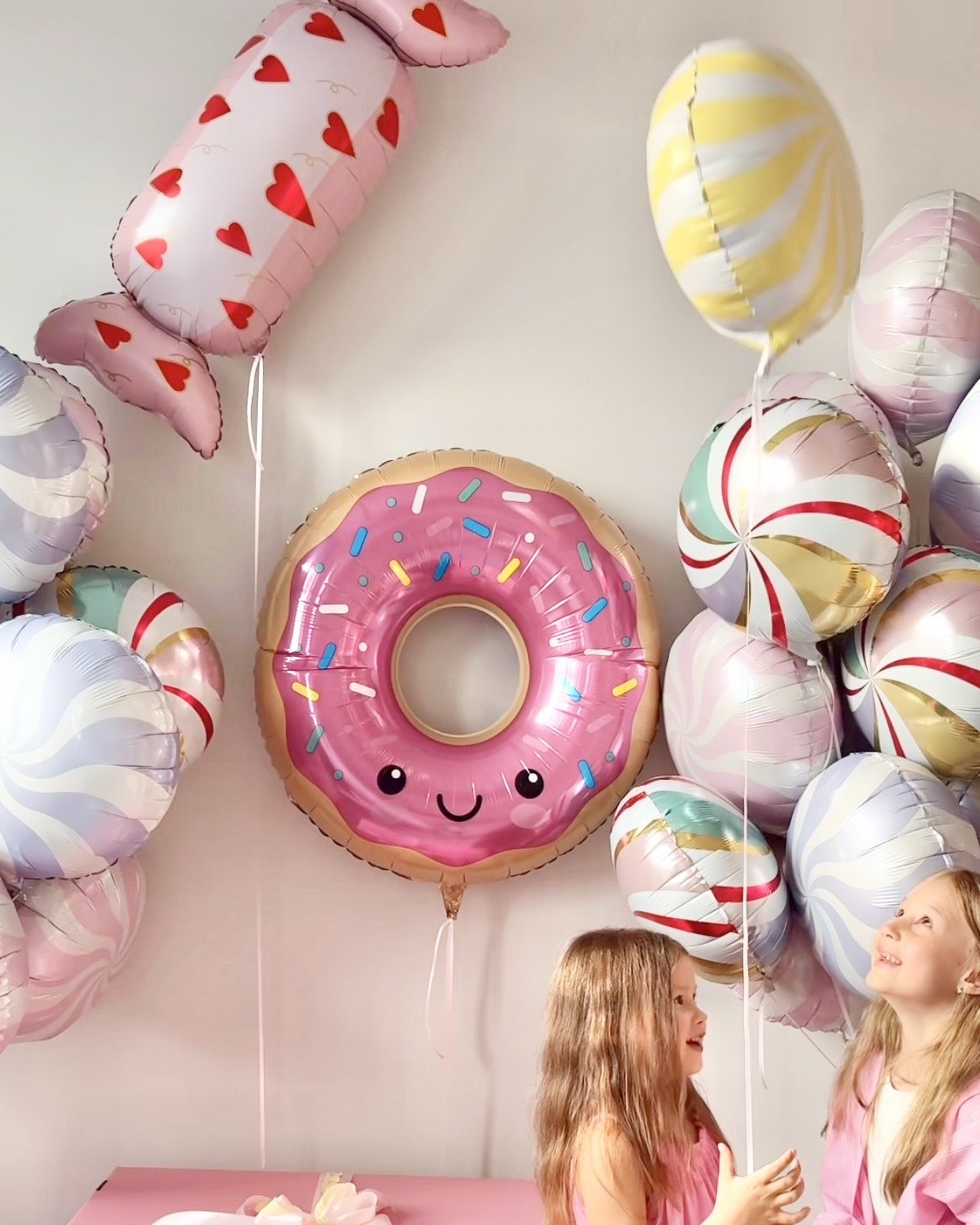 Two young girls at a birthday party with colorful helium balloons, including a large pink donut-shaped balloon with a smiling face, pink frosting, and colorful sprinkles, and various other patterned balloons in pastel colors.