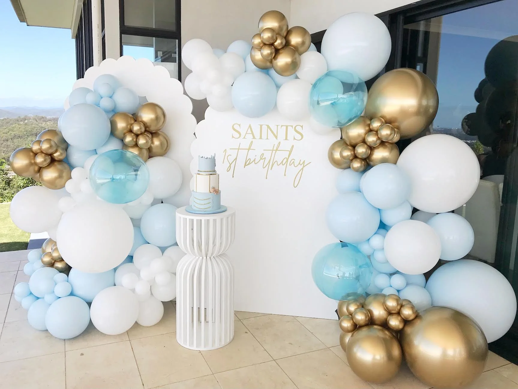 A decorative balloon display for a 1st birthday celebration with a white backdrop that has gold lettering saying "SAINTS 1st birthday." The display includes clusters of white, light blue, transparent teal, and gold balloons of various sizes, arranged