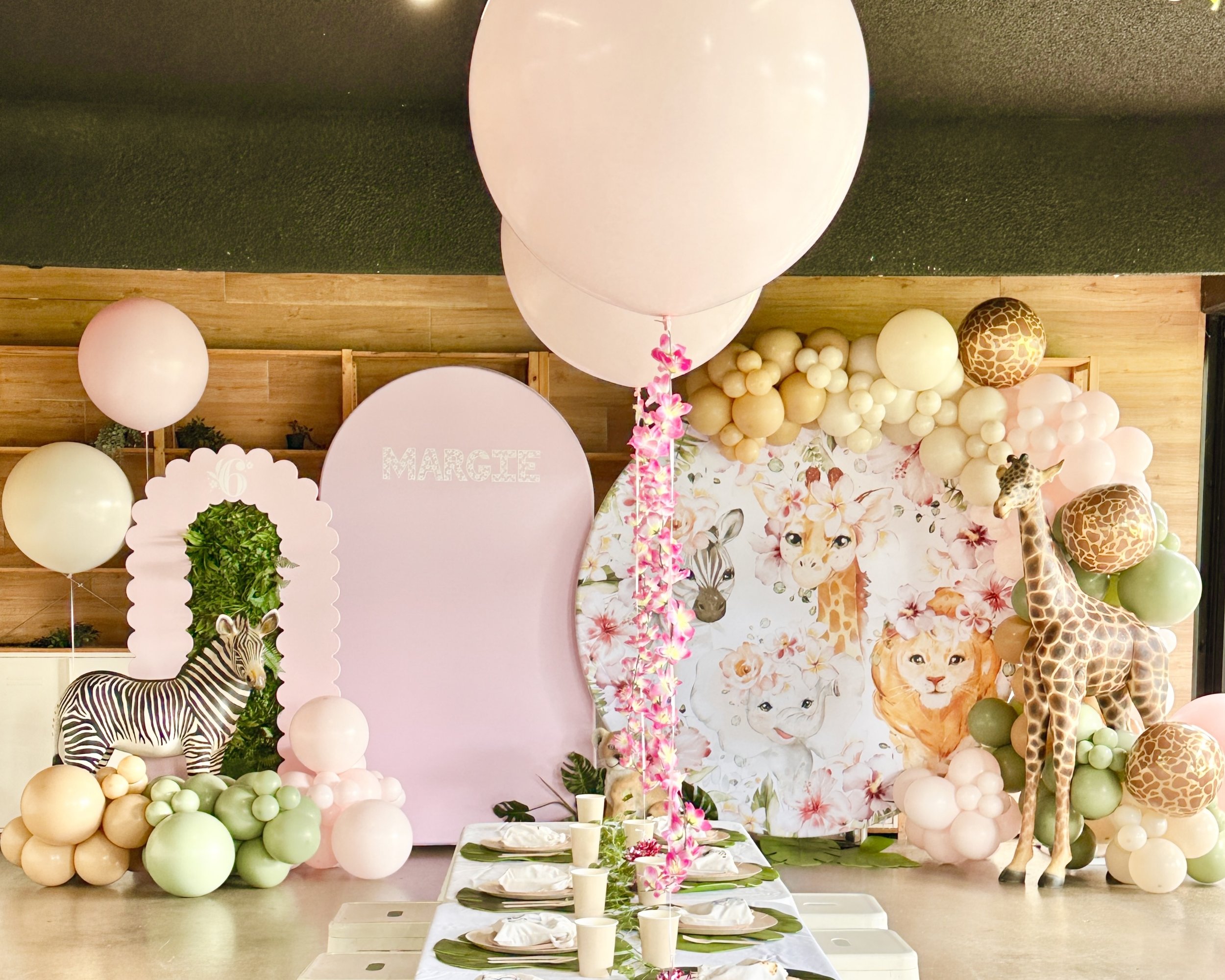 Decorative jungle animal-themed birthday party setup with balloons, plush giraffe and zebra, floral backdrop featuring lions and giraffes, and a dining table with place settings.