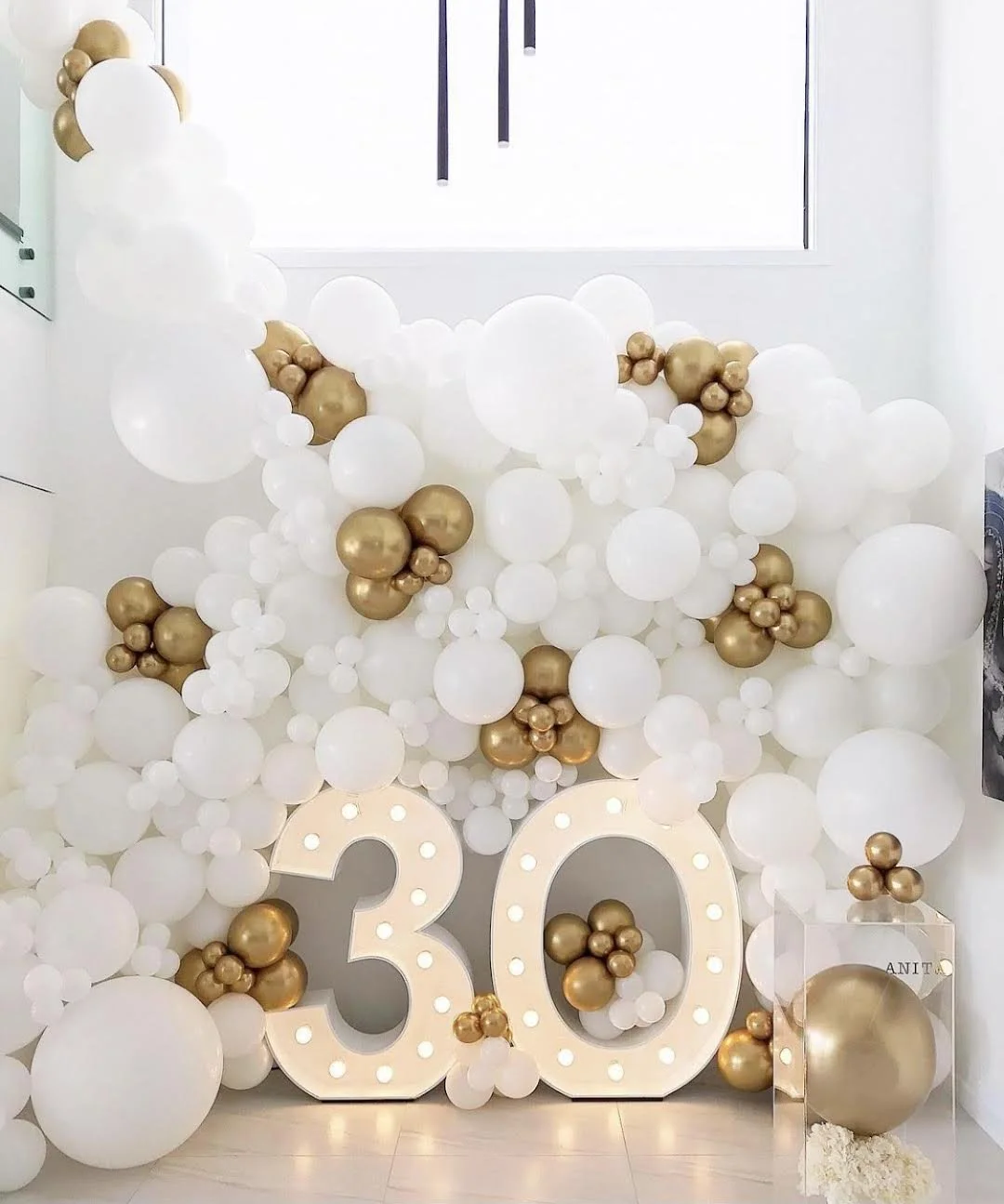 Large illuminated number 30 surrounded by white and gold balloons creating a balloon wall backdrop for a 30th birthday