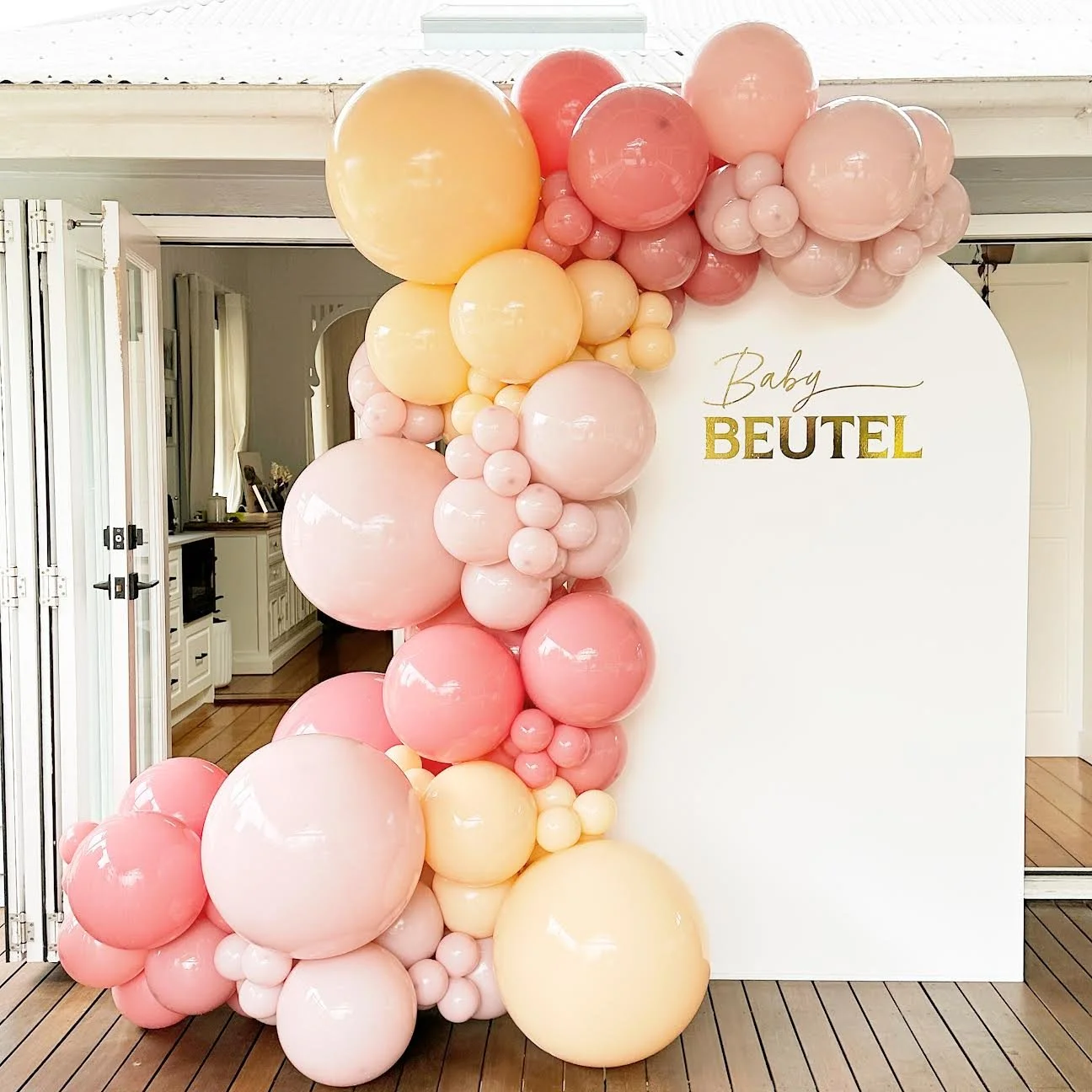 Decorative balloon arch featuring pink, peach, and cream-colored balloons next to a white backdrop with the text 'Baby BEUTEL' in gold lettering for a baby shower celebration.