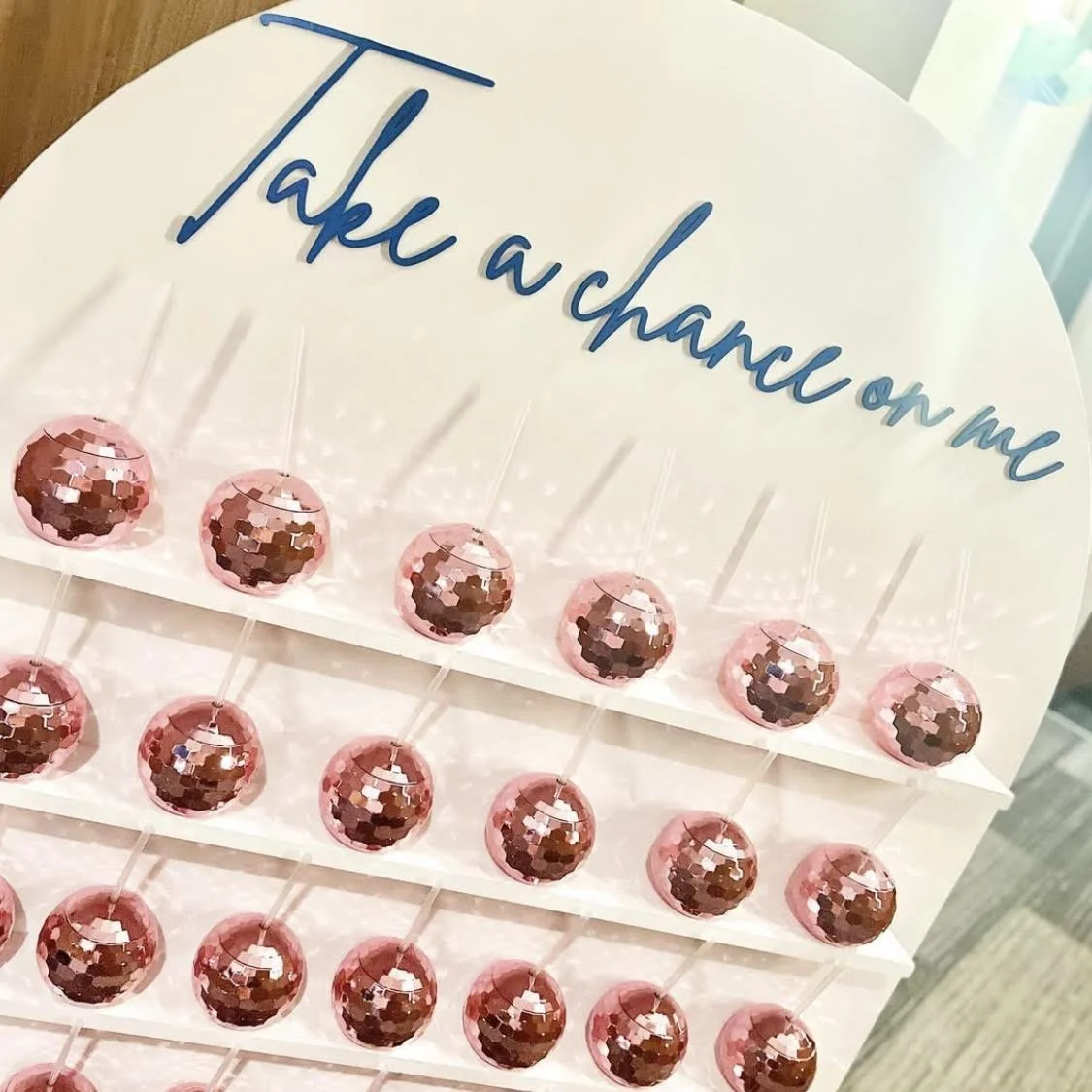 A display of pink spherical candies with reflective mosaic-like surfaces, arranged in rows on a white stand. A sign at the top reads "Take a chance on me" in blue cursive lettering.