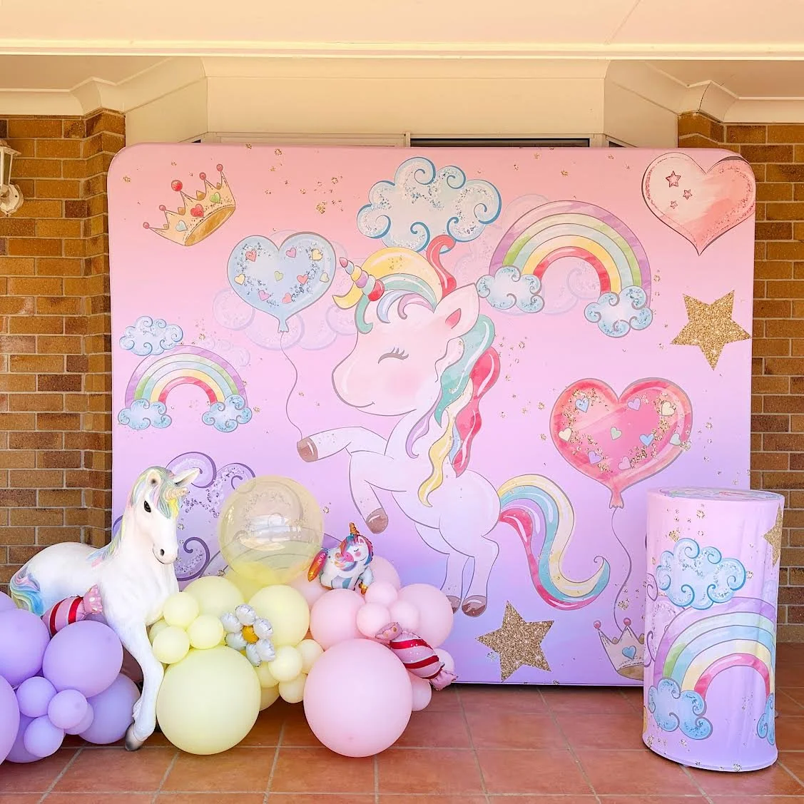 Decorative unicorn-themed birthday backdrop with balloons, featuring a pink background with rainbow, clouds, stars, hearts, and a unicorn illustration.