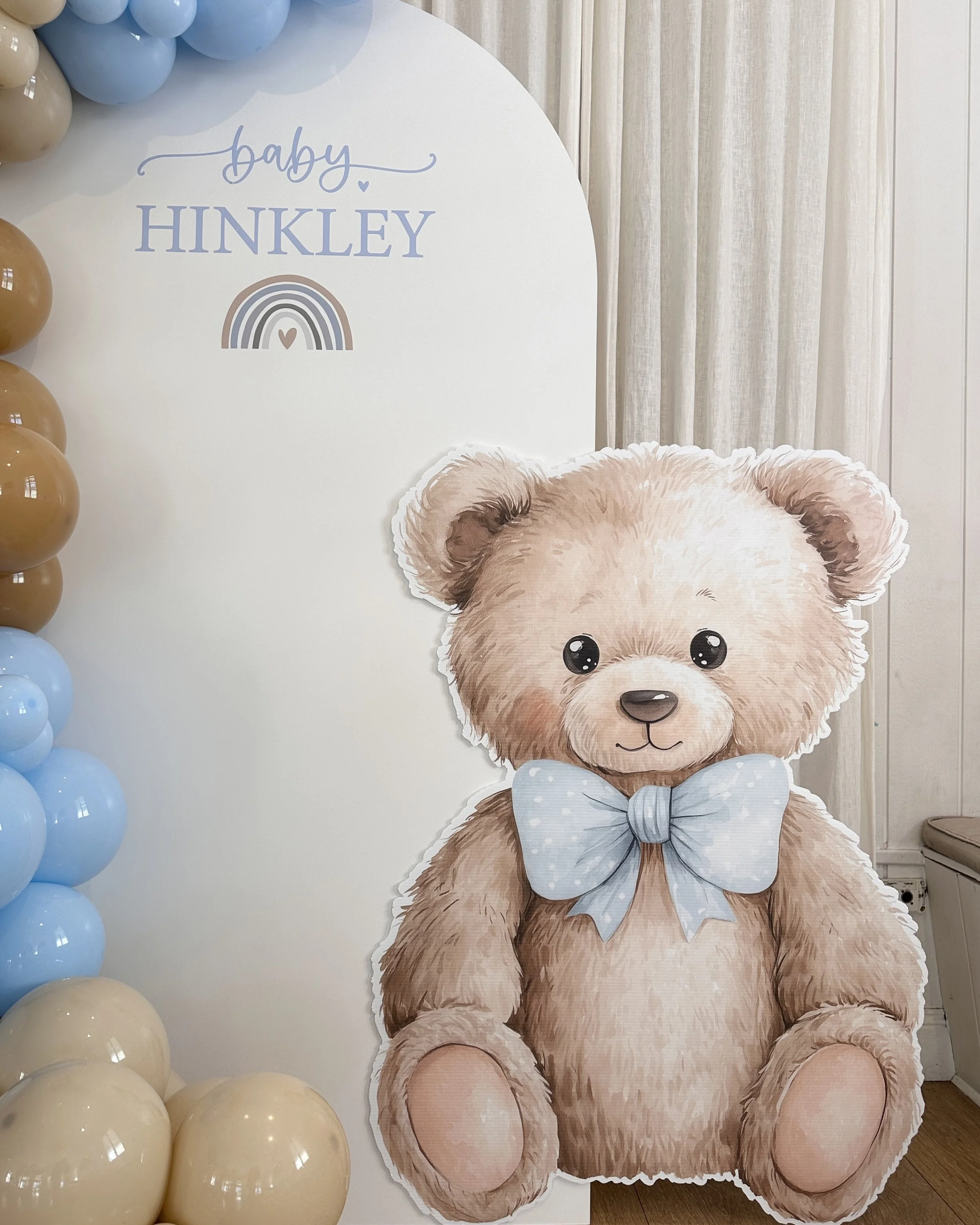 Decorative sign with the words "baby HINKLEY" above a rainbow and heart, surrounded by blue and beige balloons, and a large illustrated teddy bear with a blue ribbon.