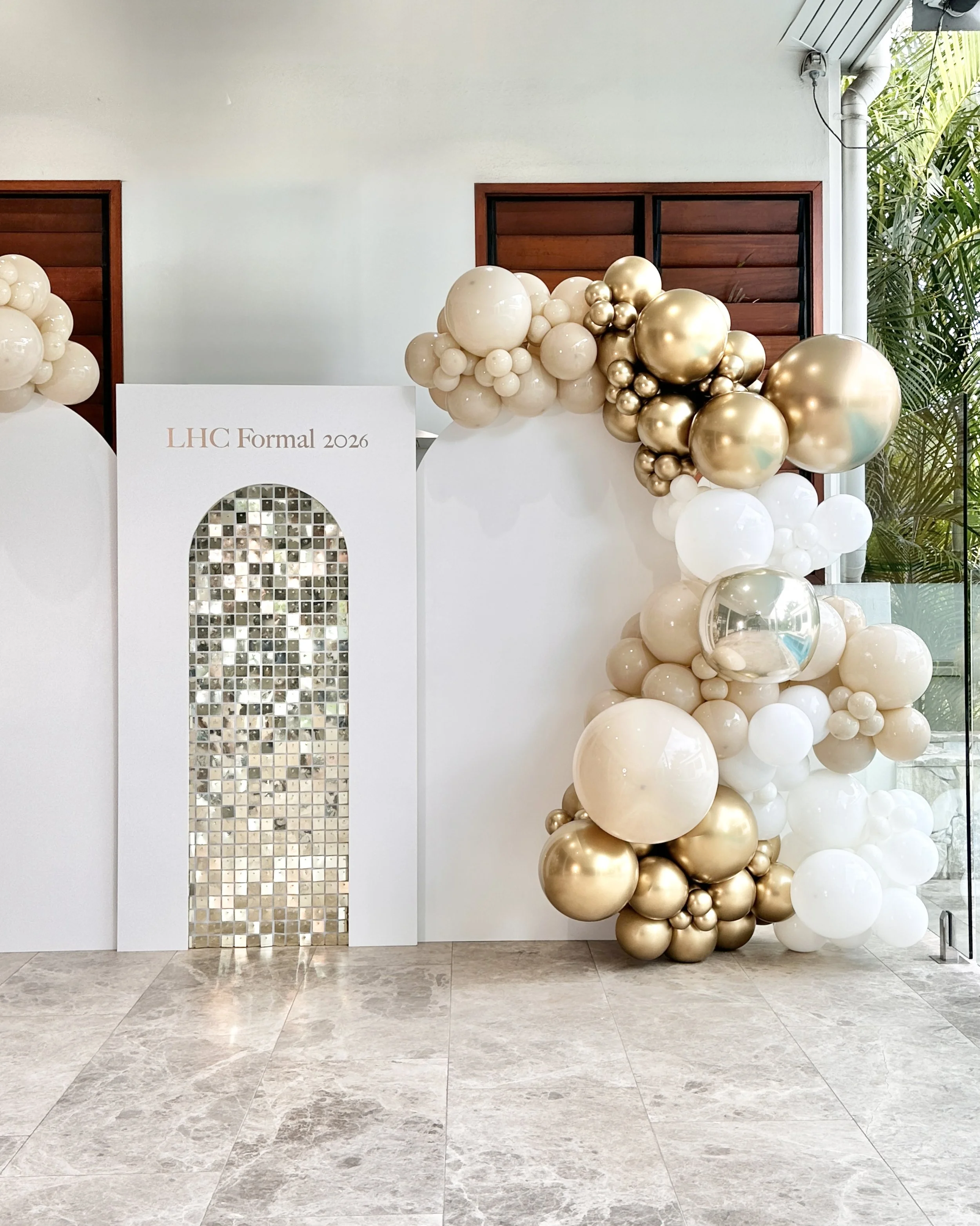 Decorative balloon arrangement with white and gold balloons against a white backdrop, with a sign reading 'LHC Formal 2026' and a gold shimmer wall.