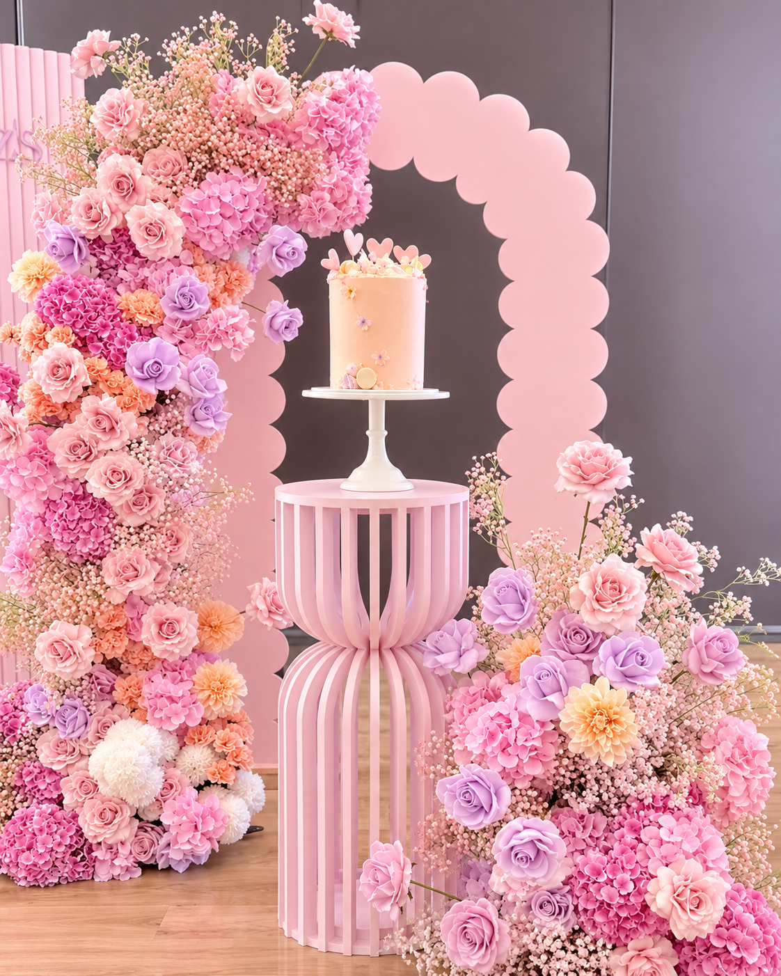A pink floral arrangement with roses and other flowers surrounds a cake on a small platform. The cake is decorated with small heart and flower designs, and is presented in an elegant display with a pink and gray backdrop.