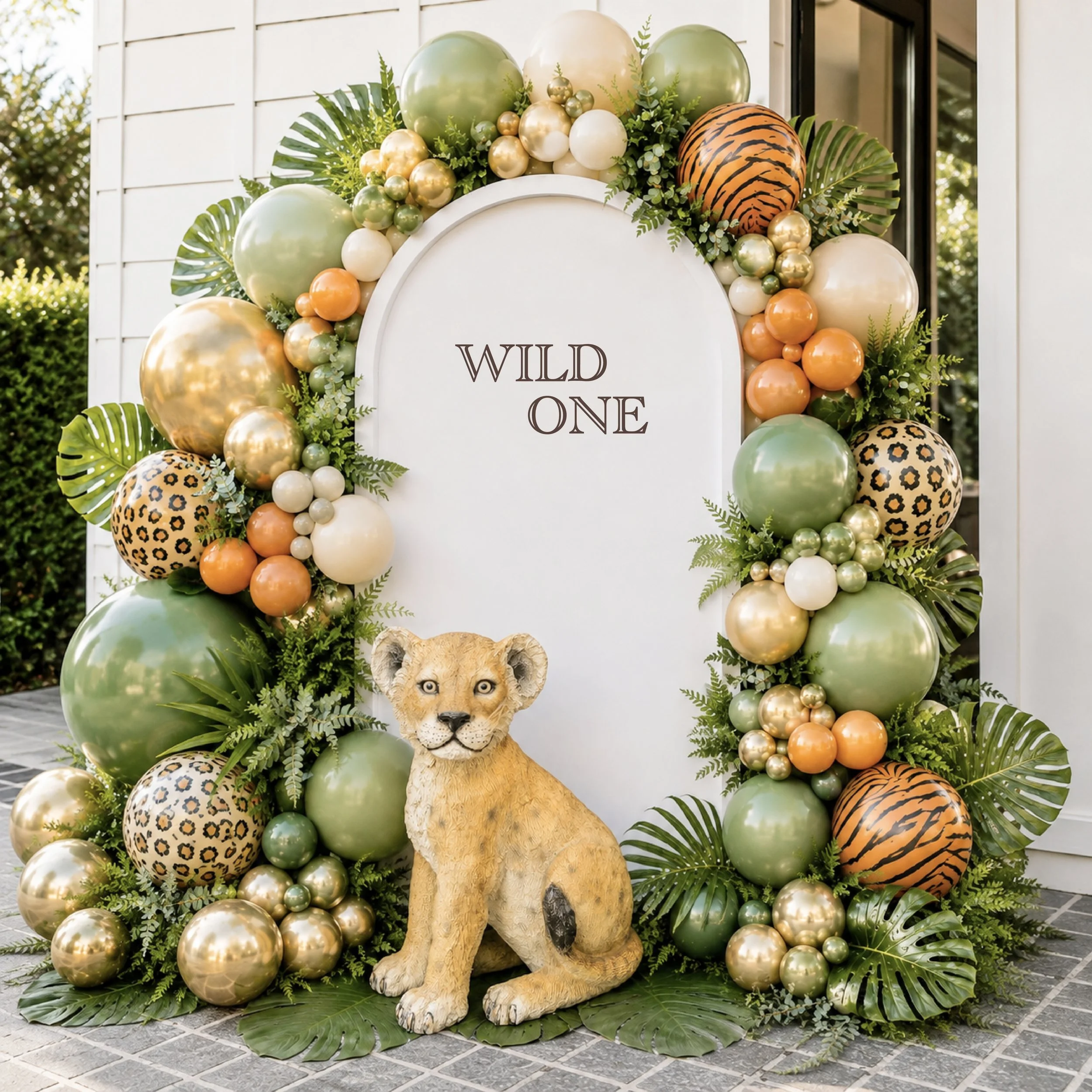 Decorative display with a white sign reading 'WILD ONE', surrounded by green, gold, ivory, orange, and leopard-print balloons, various leafy greens, and a lion statue at the bottom.