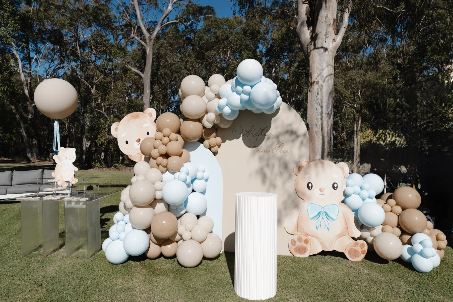 Baby shower decorations with teddy bear theme featuring a large teddy bear cutout, pastel balloons and a cake plinth