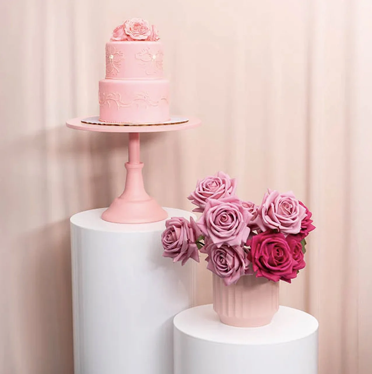 A pink, two-tiered cake with decorative pink flowers on top, displayed on a pink cake stand. Next to the cake, a pink vase holds a bouquet of pink and magenta roses. Both items are set against a light pink background.