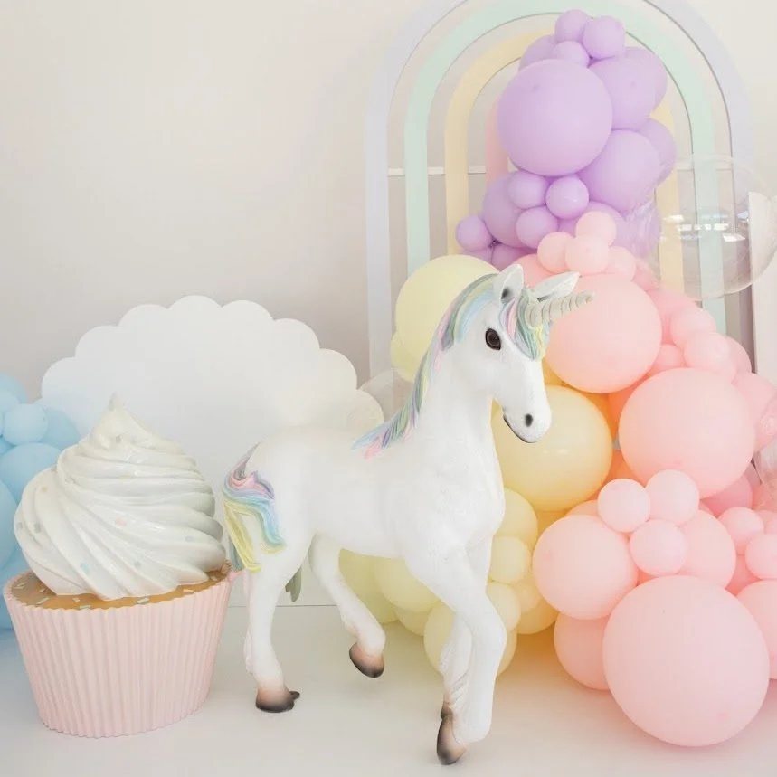 A decorative scene with a white unicorn figurine with a pastel rainbow-colored mane and tail, a giant cupcake with white frosting, and pastel-colored balloons in pink, yellow, and purple in the background.