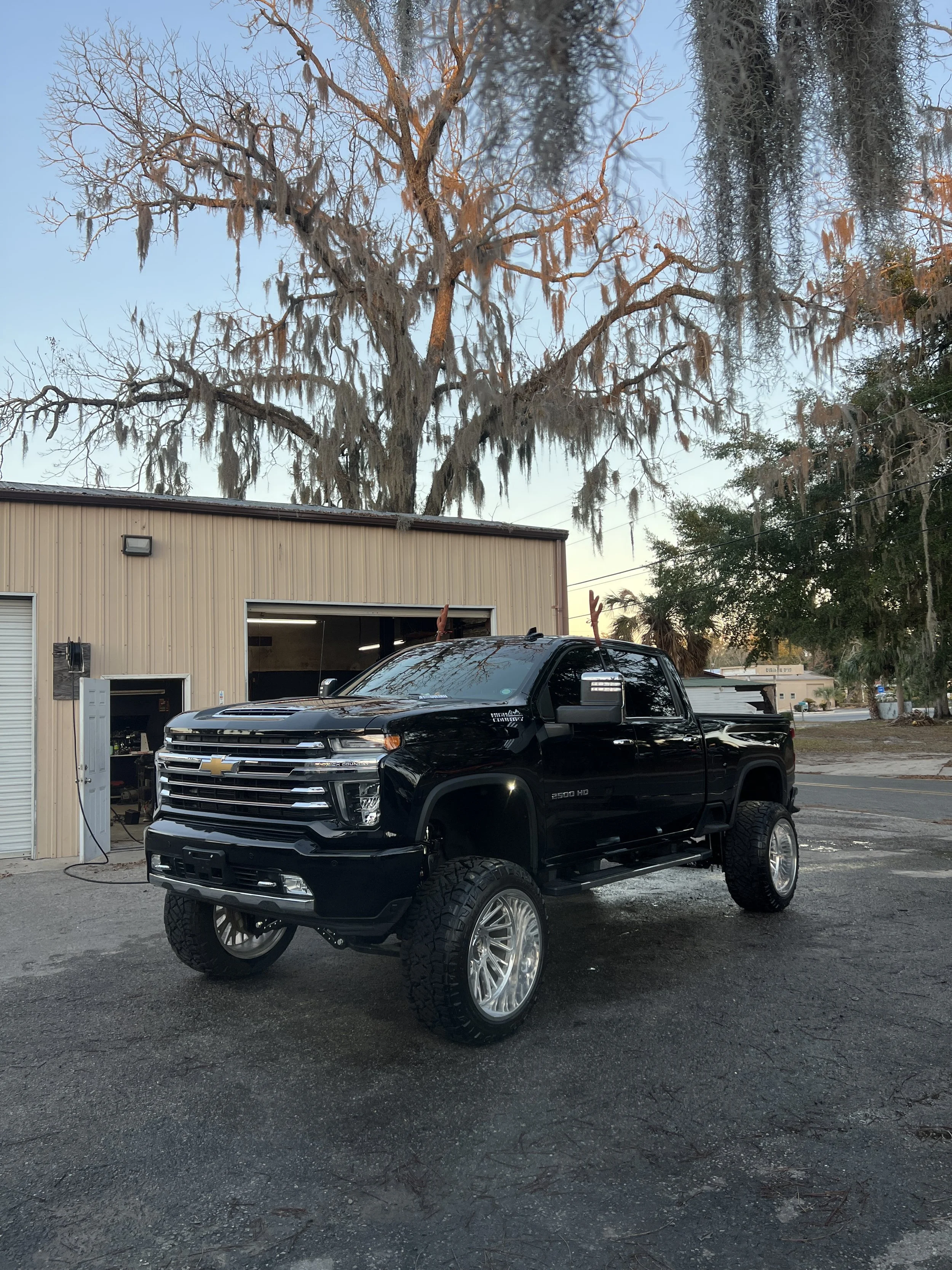 A black Chevrolet Silverado pickup truck with large tires parked outside a beige building, with a leafless tree with moss hanging from the branches and a clear sky in the background.