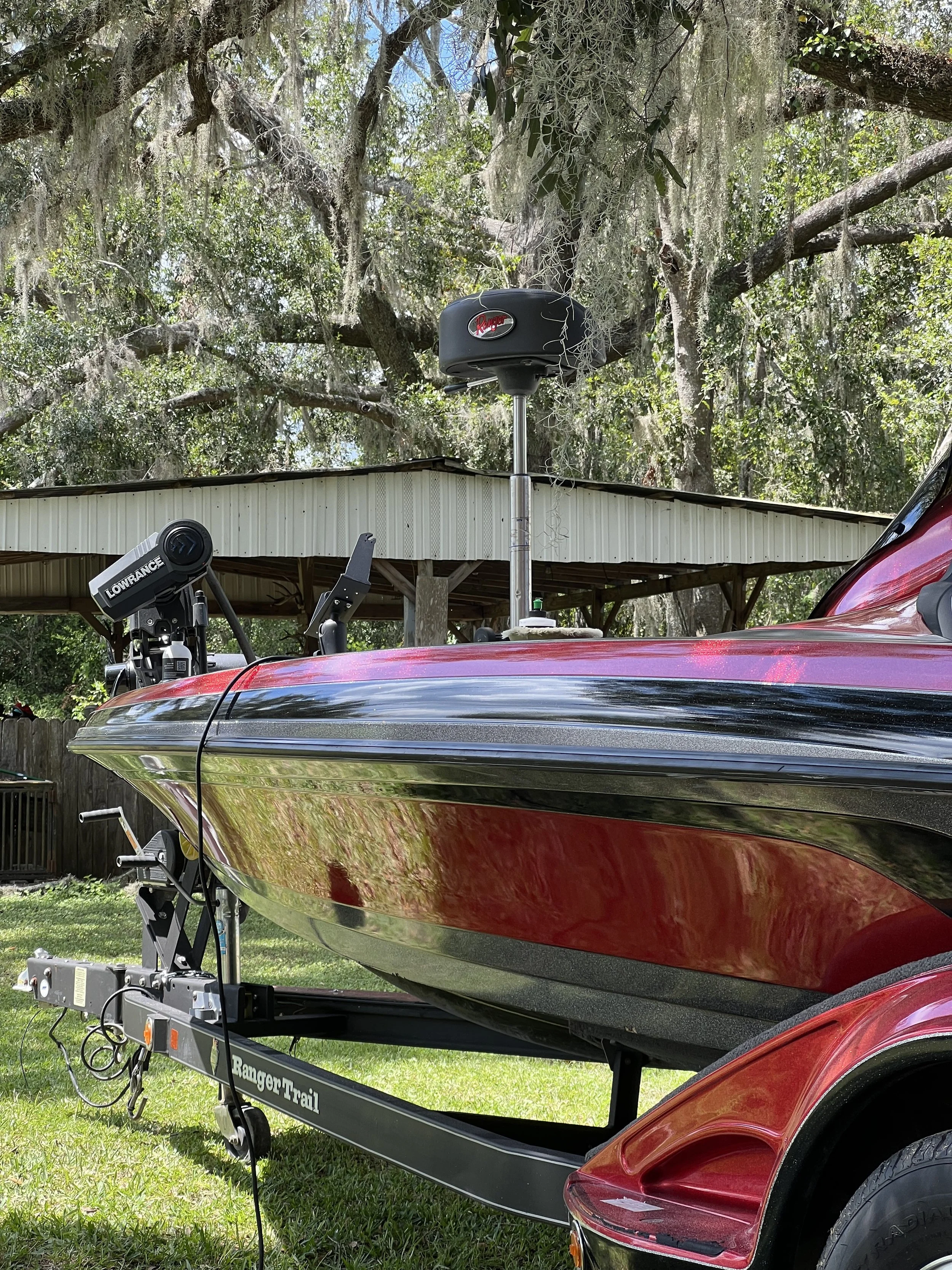 A red boat on a trailer with electronics mounted on the boat, aground in a grassy yard with trees and a shed in the background.