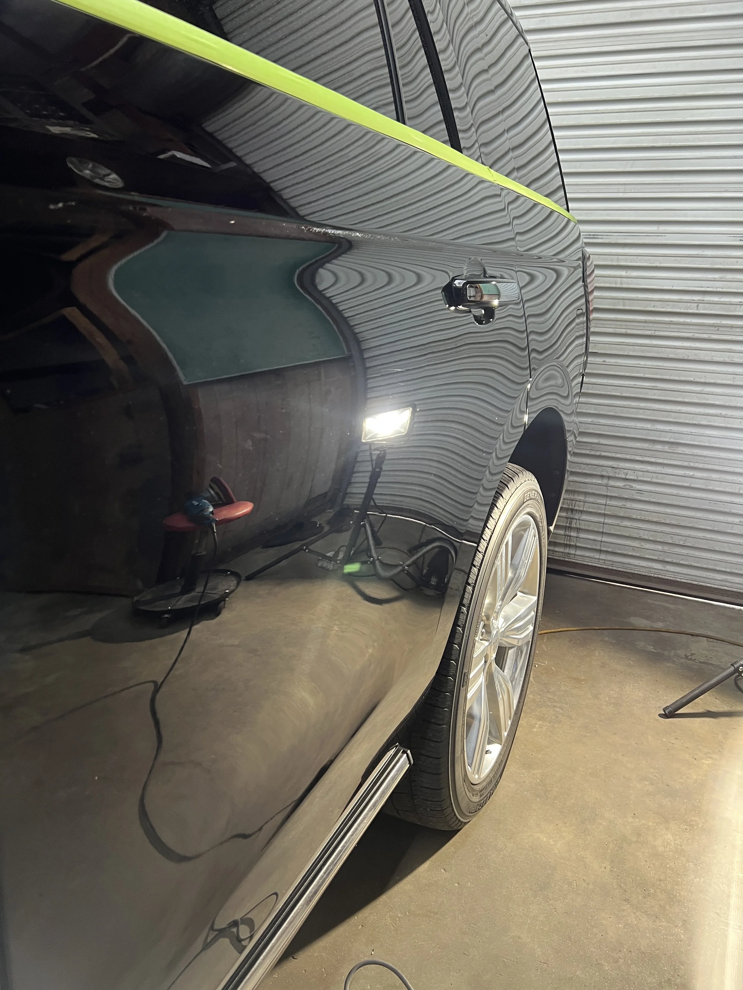 Close-up of a black car's side, showing its shiny surface, a door handle, a silver wheel, and a tire. The car is parked indoors, with a rolled metal garage door in the background. There is a yellow tape measure on the car's side, and a red stool with
