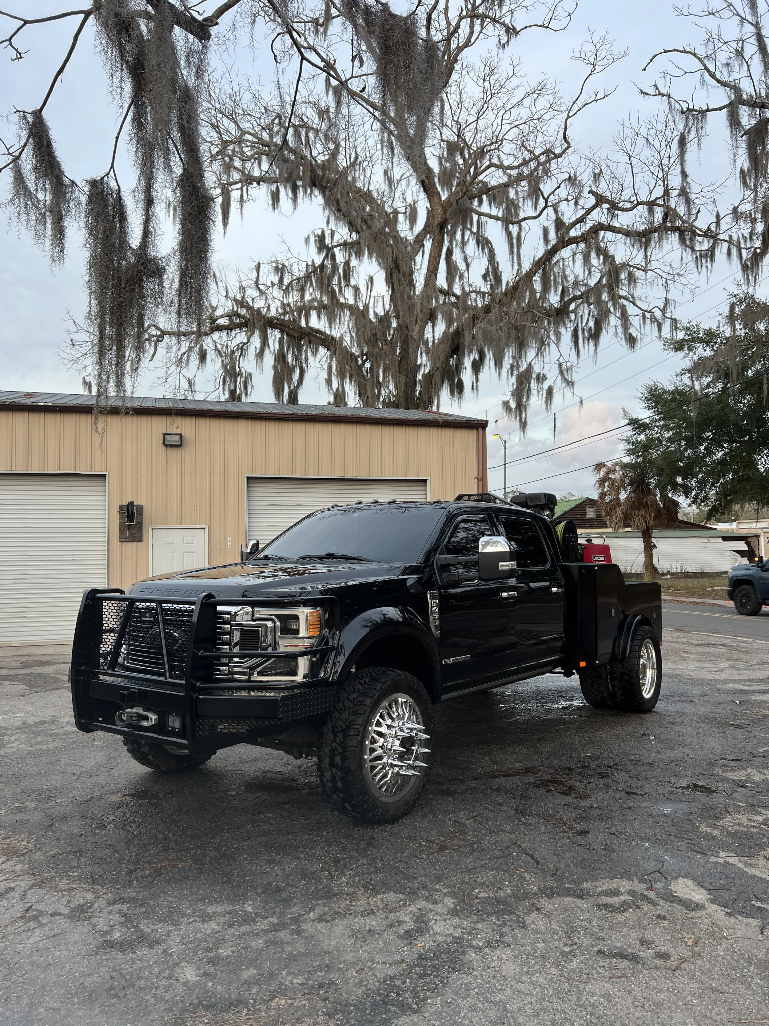 Black pickup truck with off-road tires, large chrome rims, and a heavy-duty front bumper parked on a gravel surface near a tan building with no windows and a large tree with moss hanging from its branches in the background.