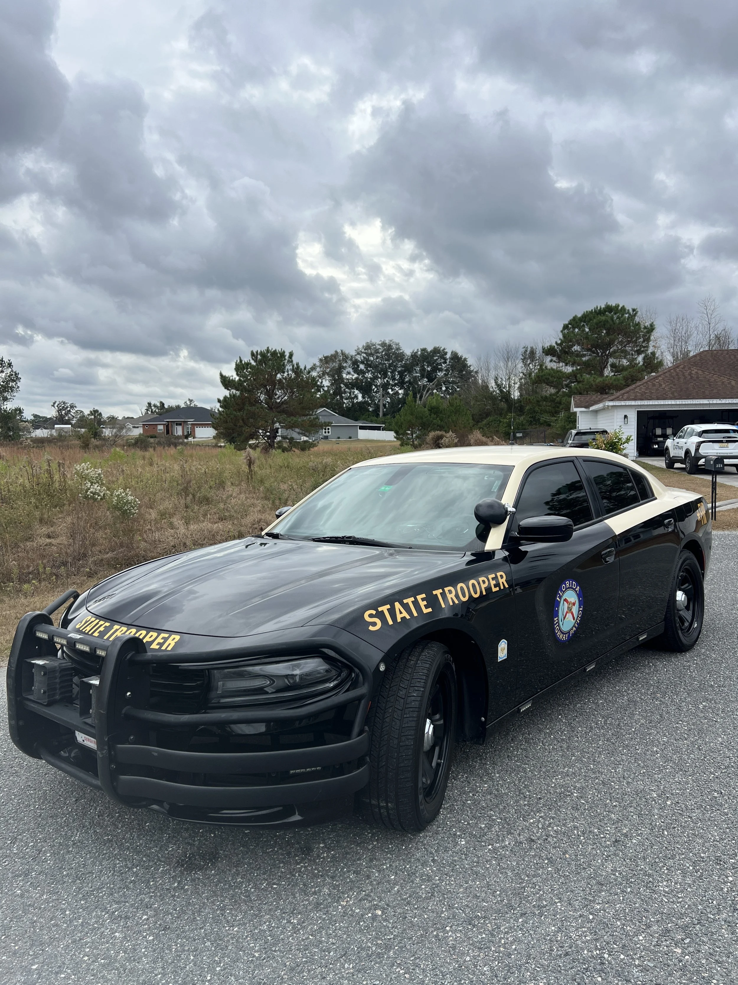 A black and white Florida State Trooper car parked on a gravel driveway under cloudy skies.
