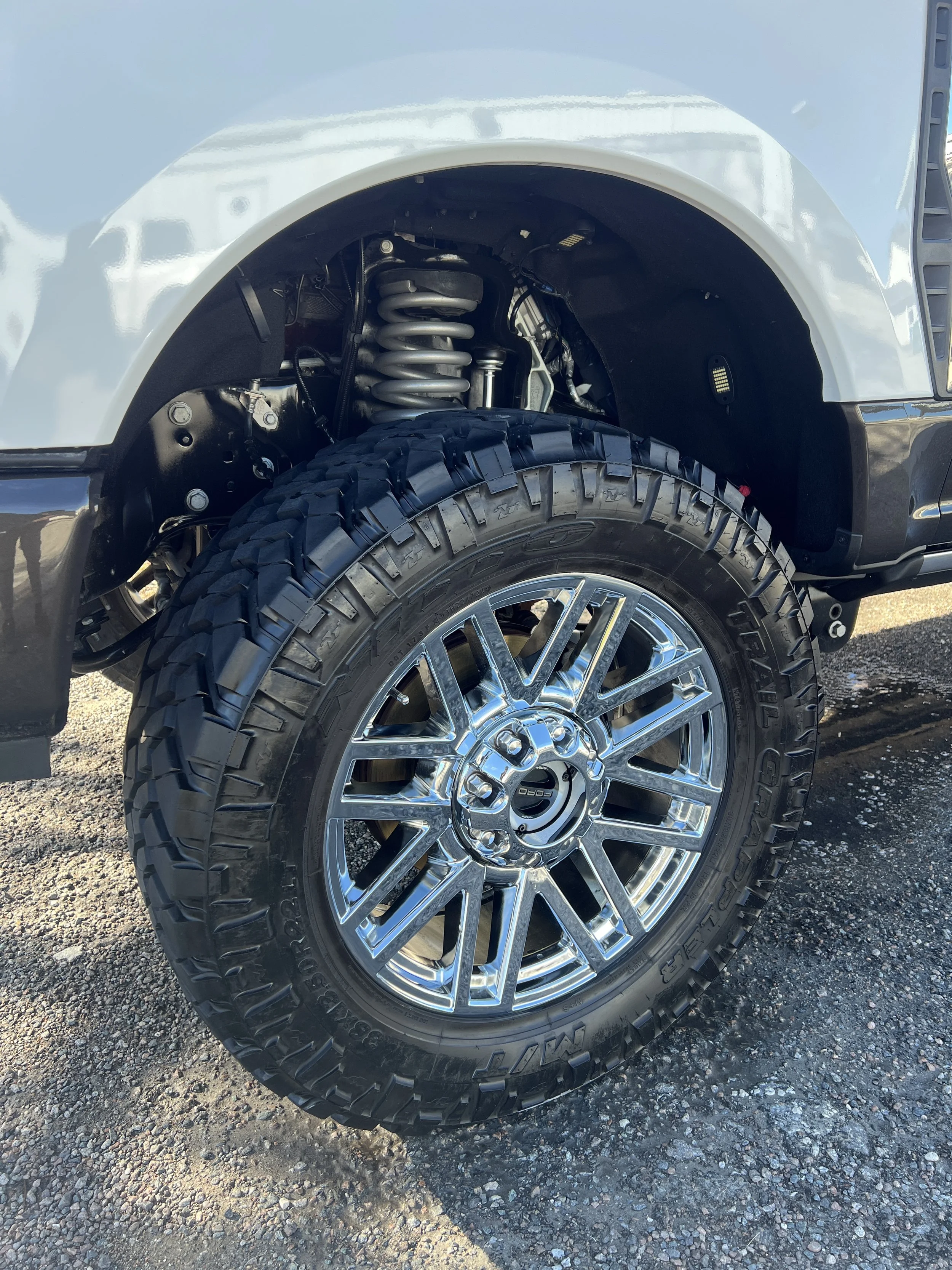 Close-up of a truck wheel with a chrome rim, mounted on a tire, with part of the open wheel well and suspension visible, outdoors on a gravel surface.
