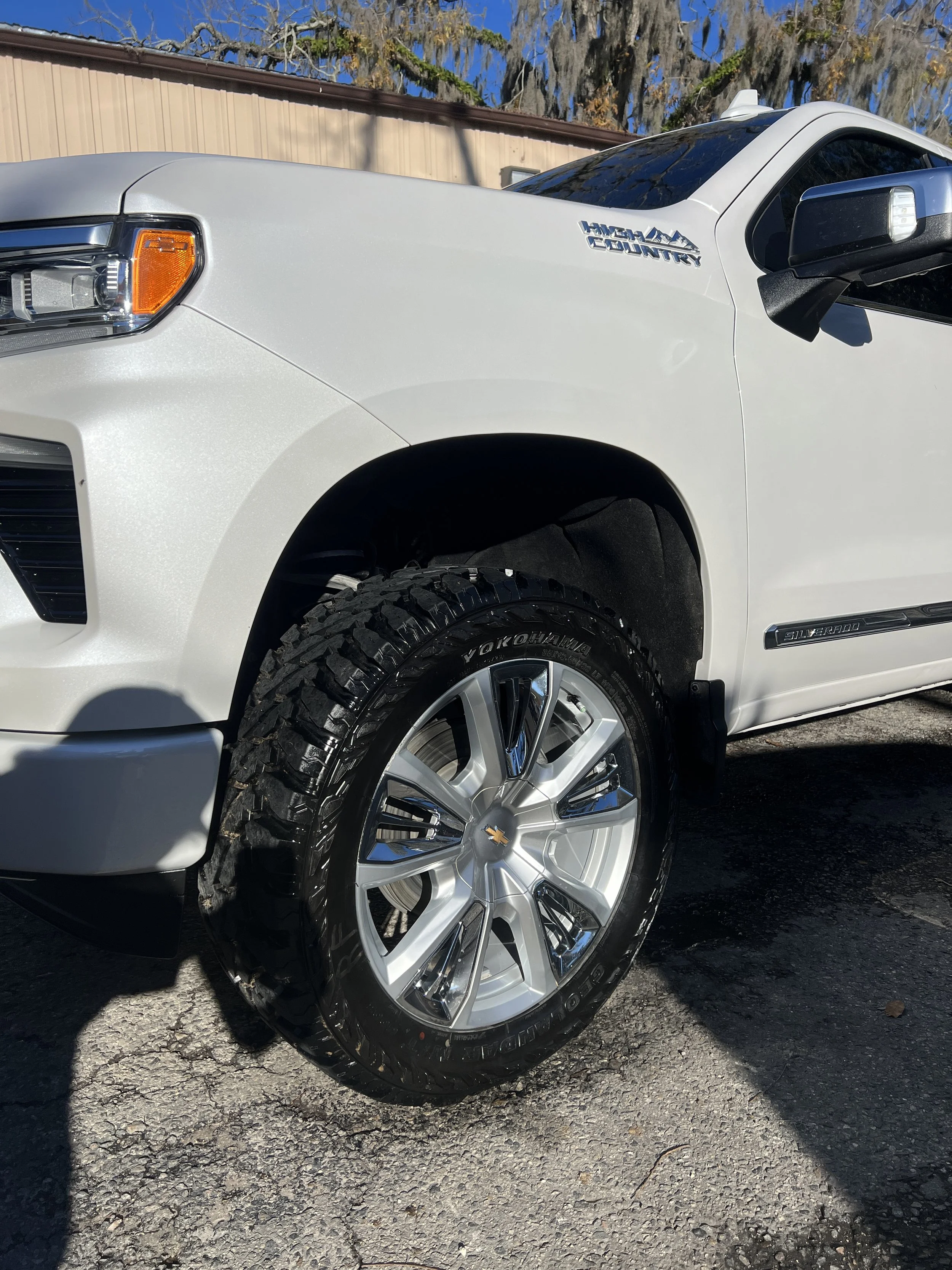 Close-up of front part of a white Chevrolet Silverado pickup truck with Off Road tires parked on asphalt, with trees and a building in the background.
